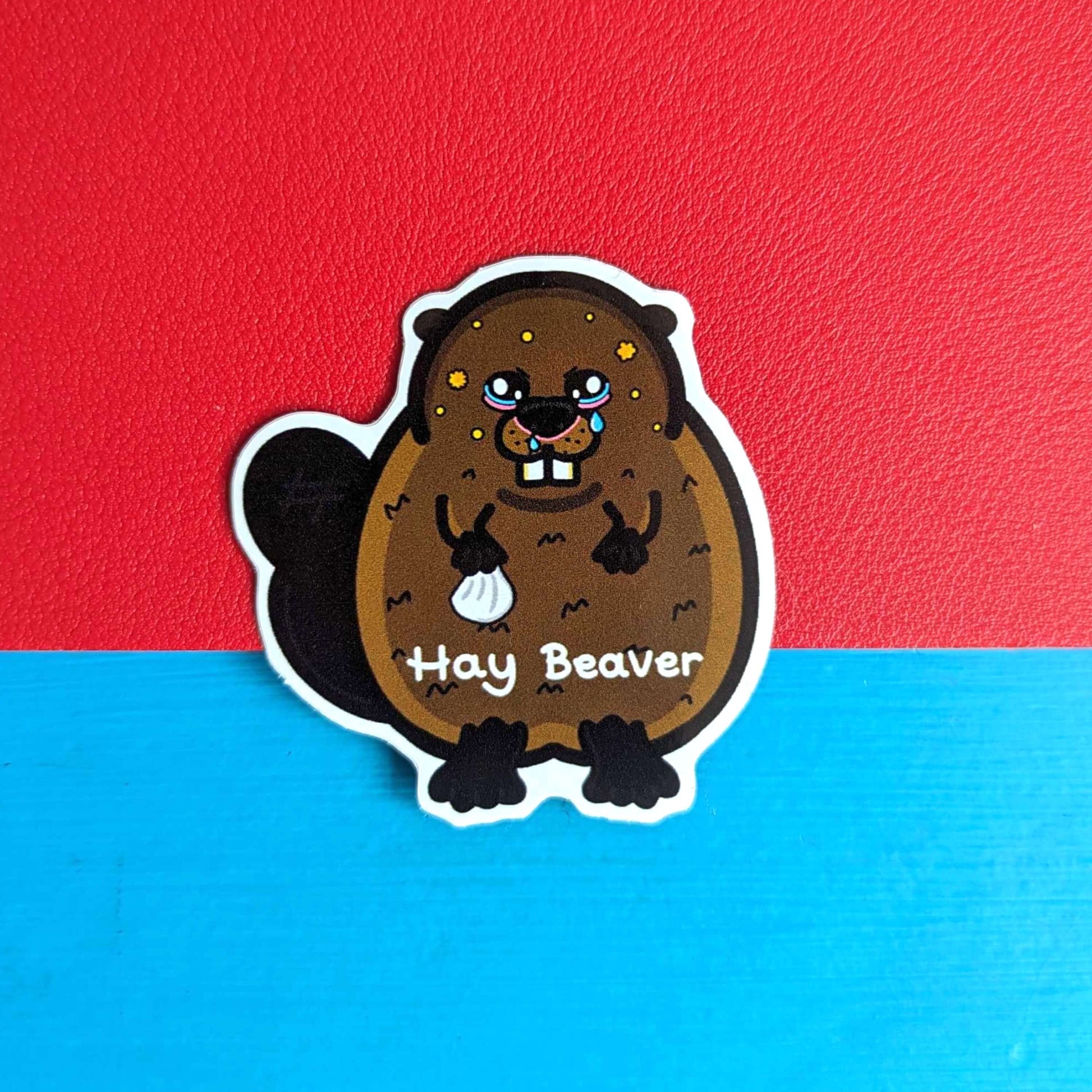 Hay Beaver Sticker on a red and blue background. The brown beaver sticker has watery eyes, dripping nose and yellow spots holding a tissue with 'hay beaver' written in white across its lower body. The hand drawn design is raising awareness for hay fever and allergic rhinitis.