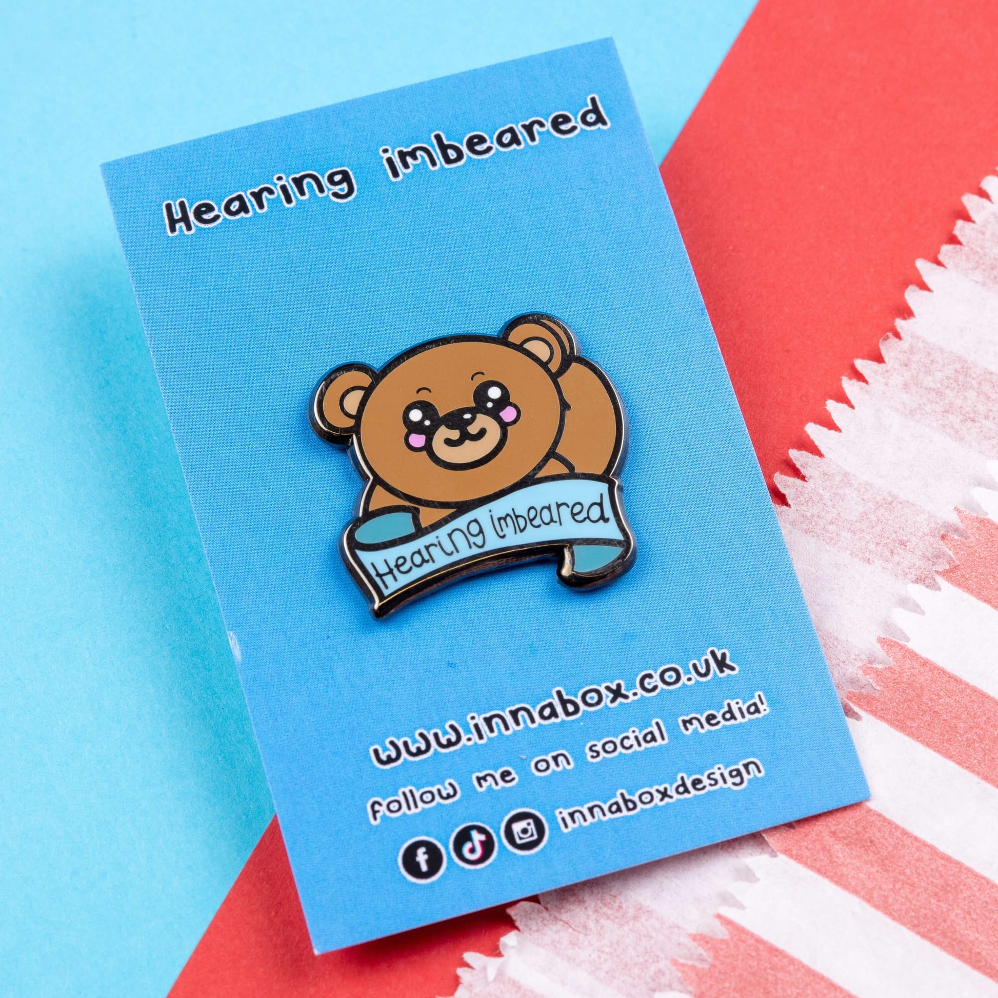 Hearing Imbeared Enamel Pin - Hearing Impaired on blue backing card on a blue and red card background. The enamel pin is a brown teddy bear with big kawaii eyes and rosy cheeks. The bear is holding one hand to its ear with a blue banner under with black text reading 'hearing imbeared'. The enamel pin is designed to raise awareness for hearing impairments, hearing loss or deafness.