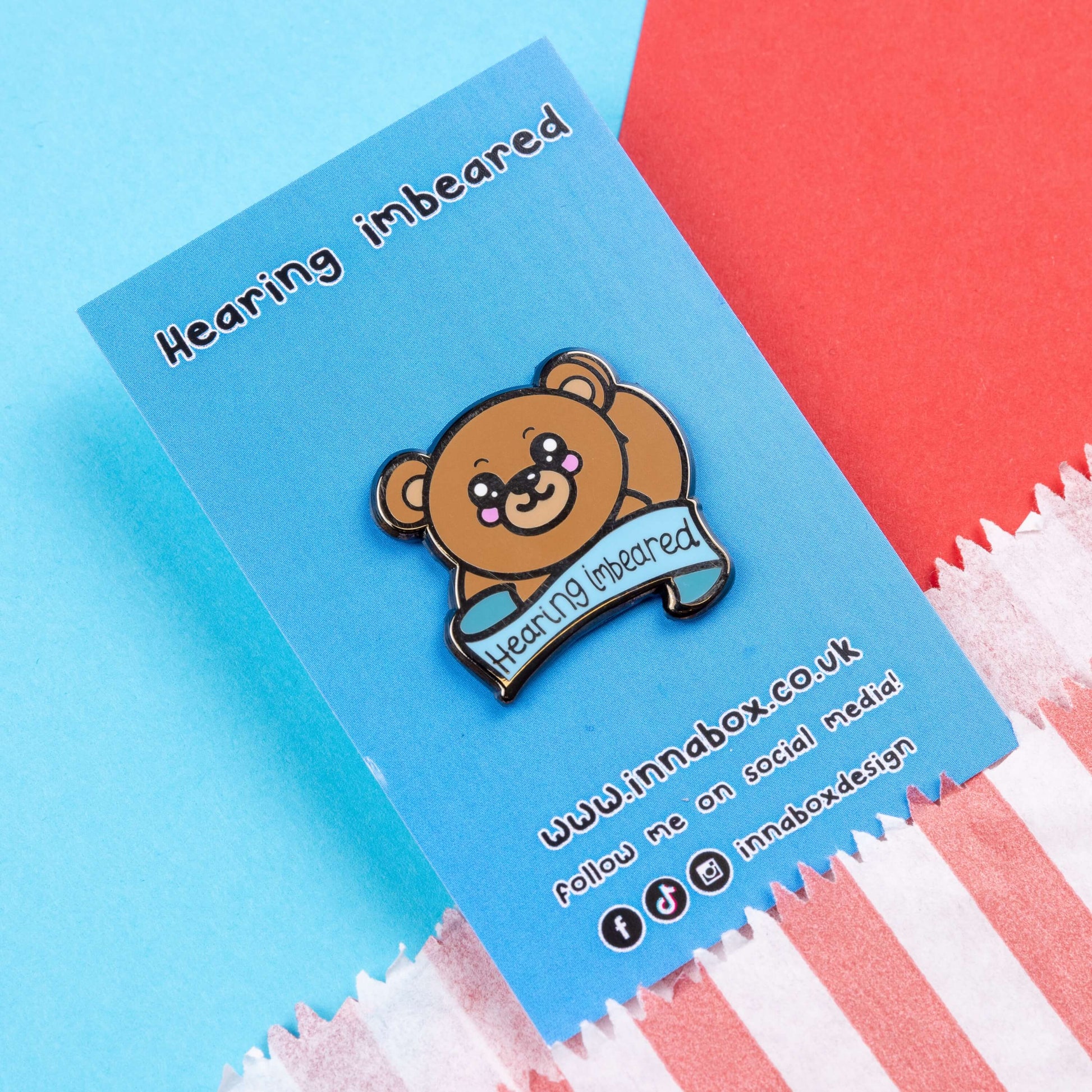 Hearing Imbeared Enamel Pin - Hearing Impaired on blue backing card on a blue and red card background. The enamel pin is a brown teddy bear with big kawaii eyes and rosy cheeks. The bear is holding one hand to its ear with a blue banner under with black text reading 'hearing imbeared'. The enamel pin is designed to raise awareness for hearing impairments, hearing loss or deafness.