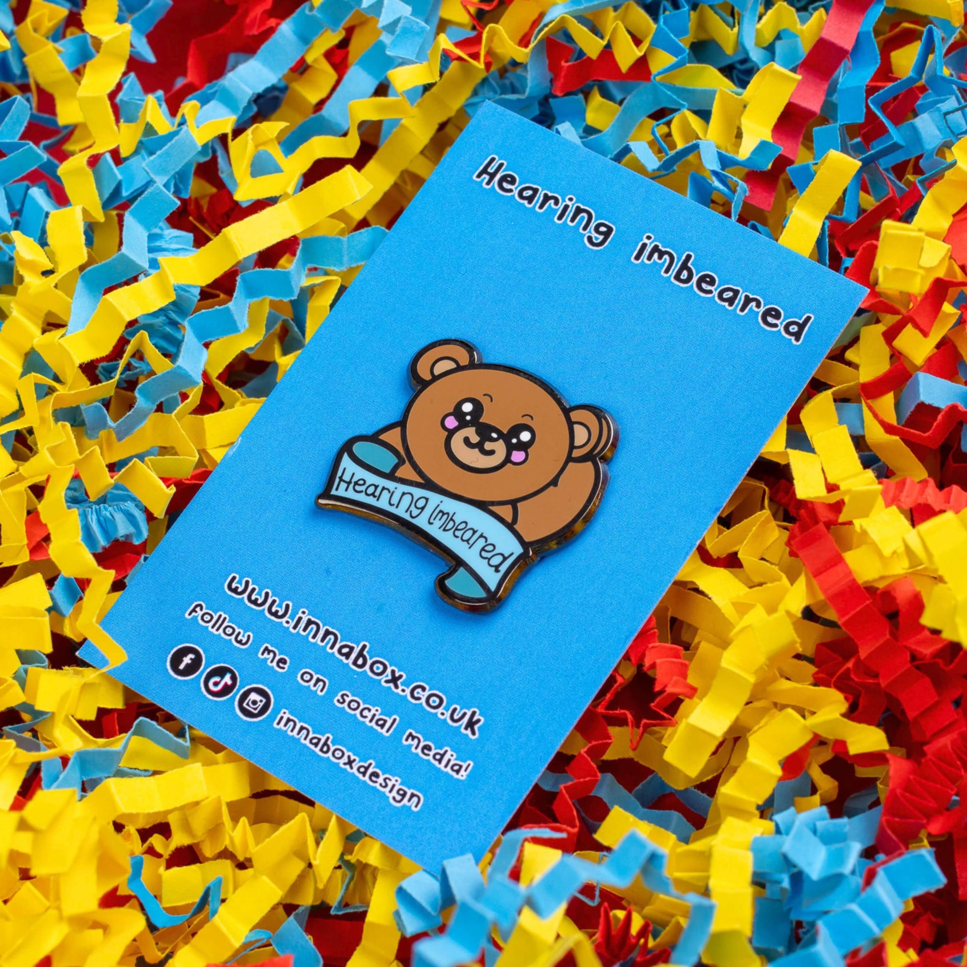 Hearing Imbeared Enamel Pin - Hearing Impaired on blue backing card on a blue, yellow and red card confetti background. The enamel pin is a brown teddy bear with big kawaii eyes and rosy cheeks. The bear is holding one hand to its ear with a blue banner under with black text reading 'hearing imbeared'. The enamel pin is designed to raise awareness for hearing impairments, hearing loss or deafness.