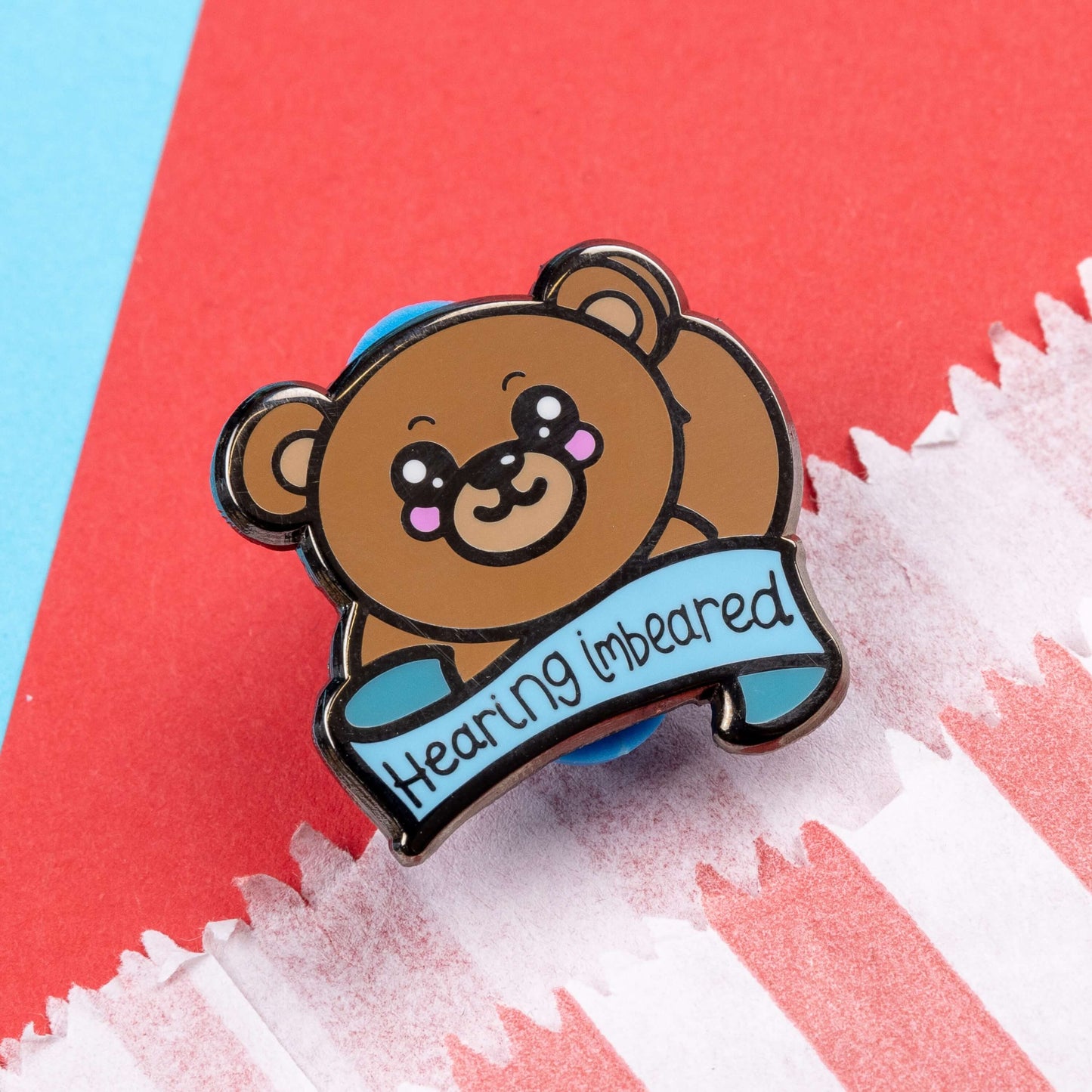 Hearing Imbeared Enamel Pin - Hearing Impaired on a blue and red card background. The enamel pin is a brown teddy bear with big kawaii eyes and rosy cheeks. The bear is holding one hand to its ear with a blue banner under with black text reading 'hearing imbeared'. The enamel pin is designed to raise awareness for hearing impairments, hearing loss or deafness.