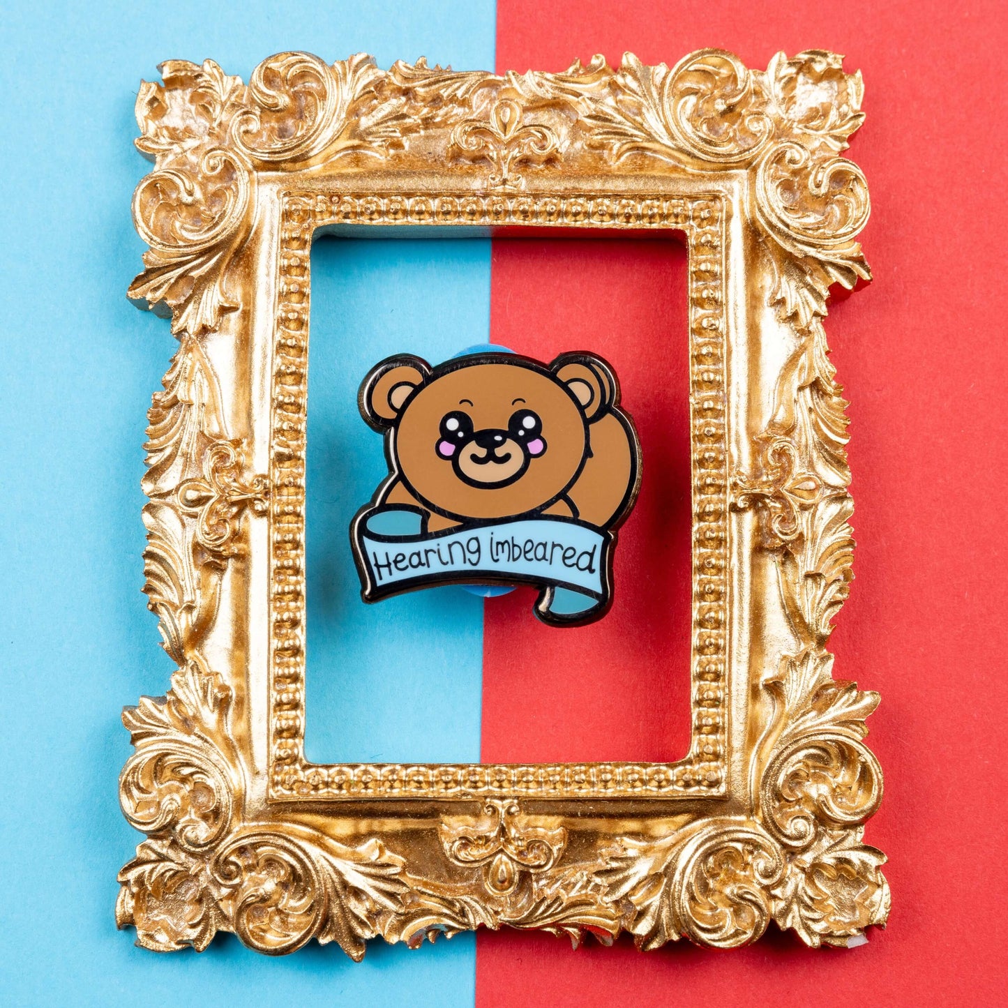 Hearing Imbeared Enamel Pin - Hearing Impaired on a blue and red card background inside a gold ornate frame. The enamel pin is a brown teddy bear with big kawaii eyes and rosy cheeks. The bear is holding one hand to its ear with a blue banner under with black text reading 'hearing imbeared'. The enamel pin is designed to raise awareness for hearing impairments, hearing loss or deafness.