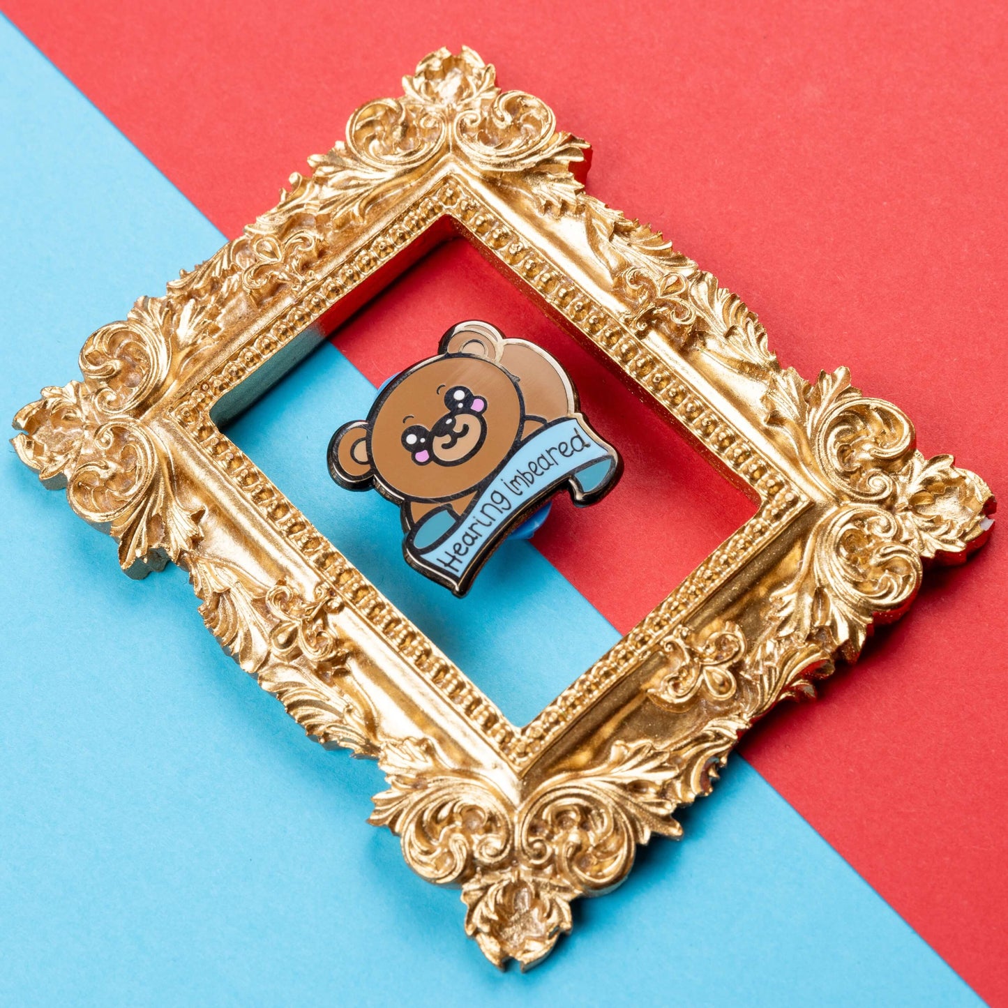 Hearing Imbeared Enamel Pin - Hearing Impaired on a blue and red card background inside a gold ornate frame. The enamel pin is a brown teddy bear with big kawaii eyes and rosy cheeks. The bear is holding one hand to its ear with a blue banner under with black text reading 'hearing imbeared'. The enamel pin is designed to raise awareness for hearing impairments, hearing loss or deafness.