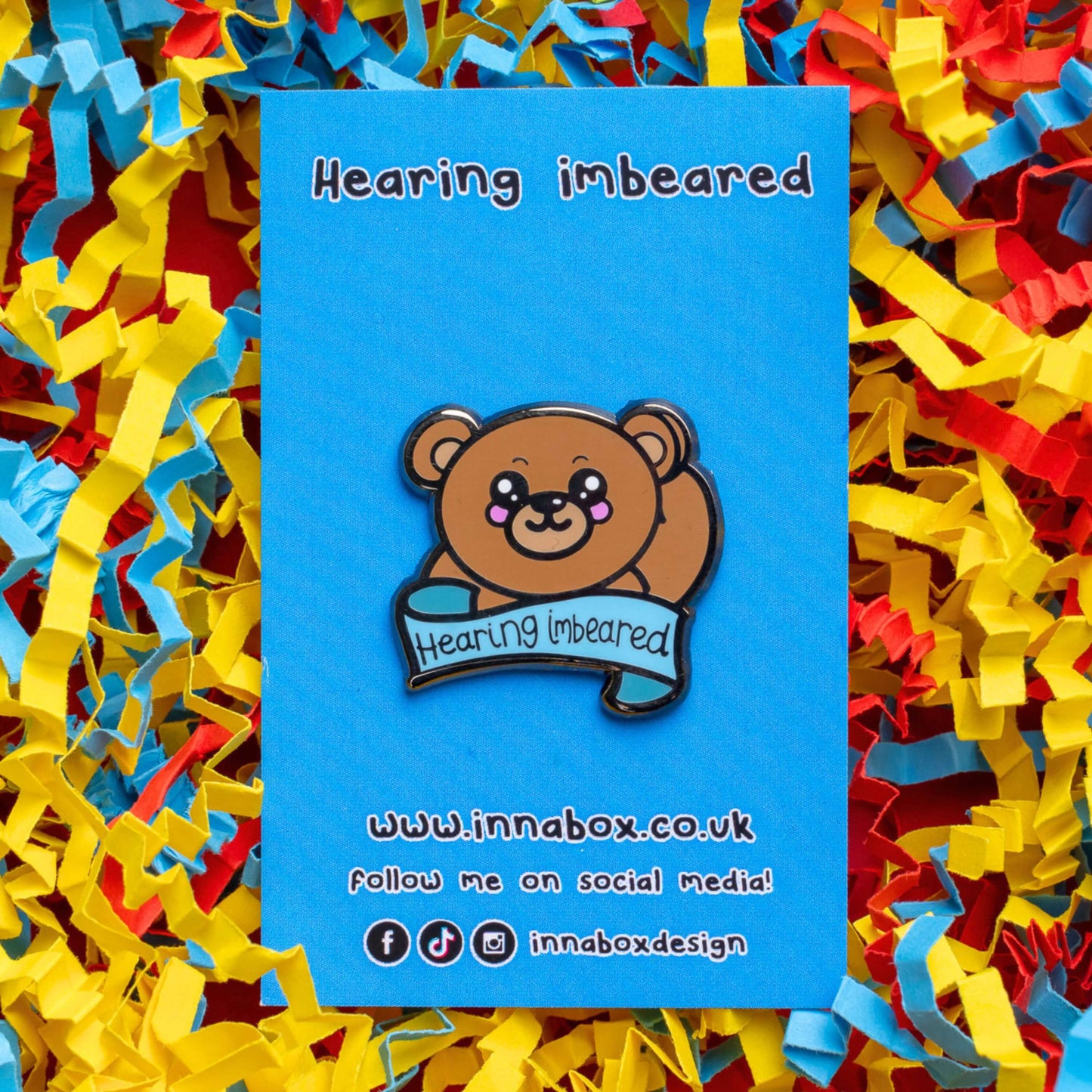 Hearing Imbeared Enamel Pin - Hearing Impaired on blue backing card on a blue, yellow and red card confetti background. The enamel pin is a brown teddy bear with big kawaii eyes and rosy cheeks. The bear is holding one hand to its ear with a blue banner under with black text reading 'hearing imbeared'. The enamel pin is designed to raise awareness for hearing impairments, hearing loss or deafness.