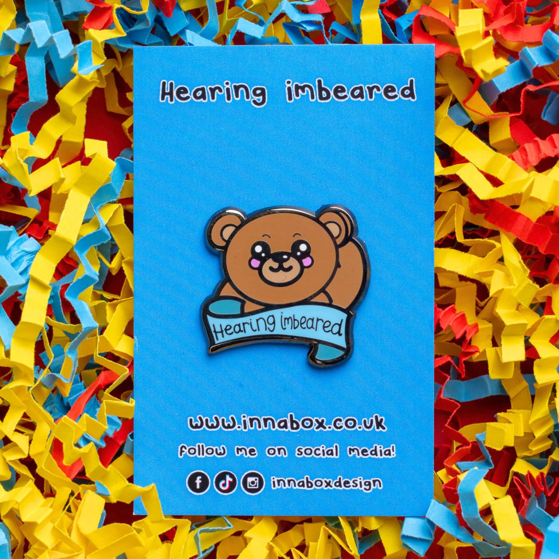 Hearing Imbeared Enamel Pin - Hearing Impaired on blue backing card on a blue, yellow and red card confetti background. The enamel pin is a brown teddy bear with big kawaii eyes and rosy cheeks. The bear is holding one hand to its ear with a blue banner under with black text reading 'hearing imbeared'. The enamel pin is designed to raise awareness for hearing impairments, hearing loss or deafness.