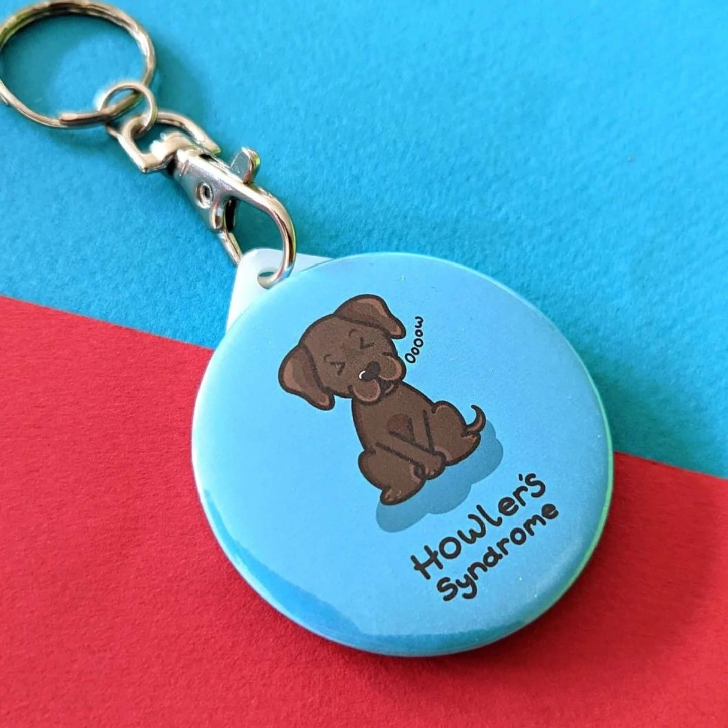 Howlers Syndrome Keyring - Fowlers Syndrome shown on a red and blue background. The circular blue keyring has an illustration of a brown dog with a pained expression and paws over its bladder. 'Howler's Syndrome' is written underneath the dog in black text. The hand drawn design is made to raise awareness for Fowlers Syndrome.