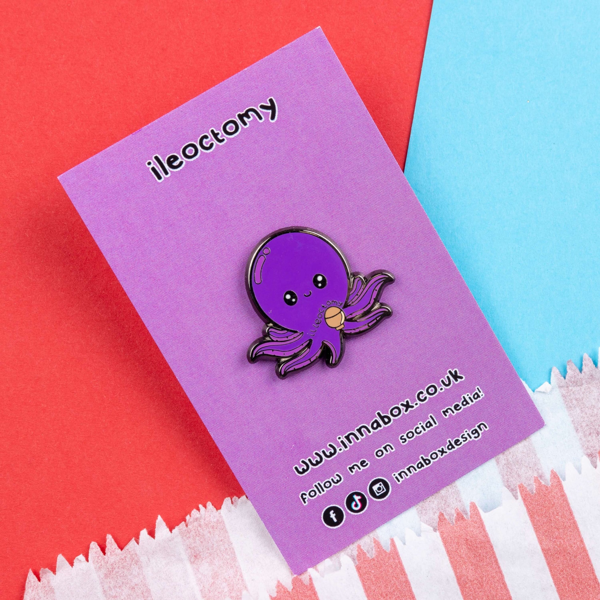 Ileoctomy Enamel Pin - Ileostomy on purple backing card laid on a blue and red card background. The enamel pin is a cute smiling purple octopus sticker with text saying ileoctomy on its belly with a stoma bag underneath. Enamel pin designed to raise awareness for Ileostomy.