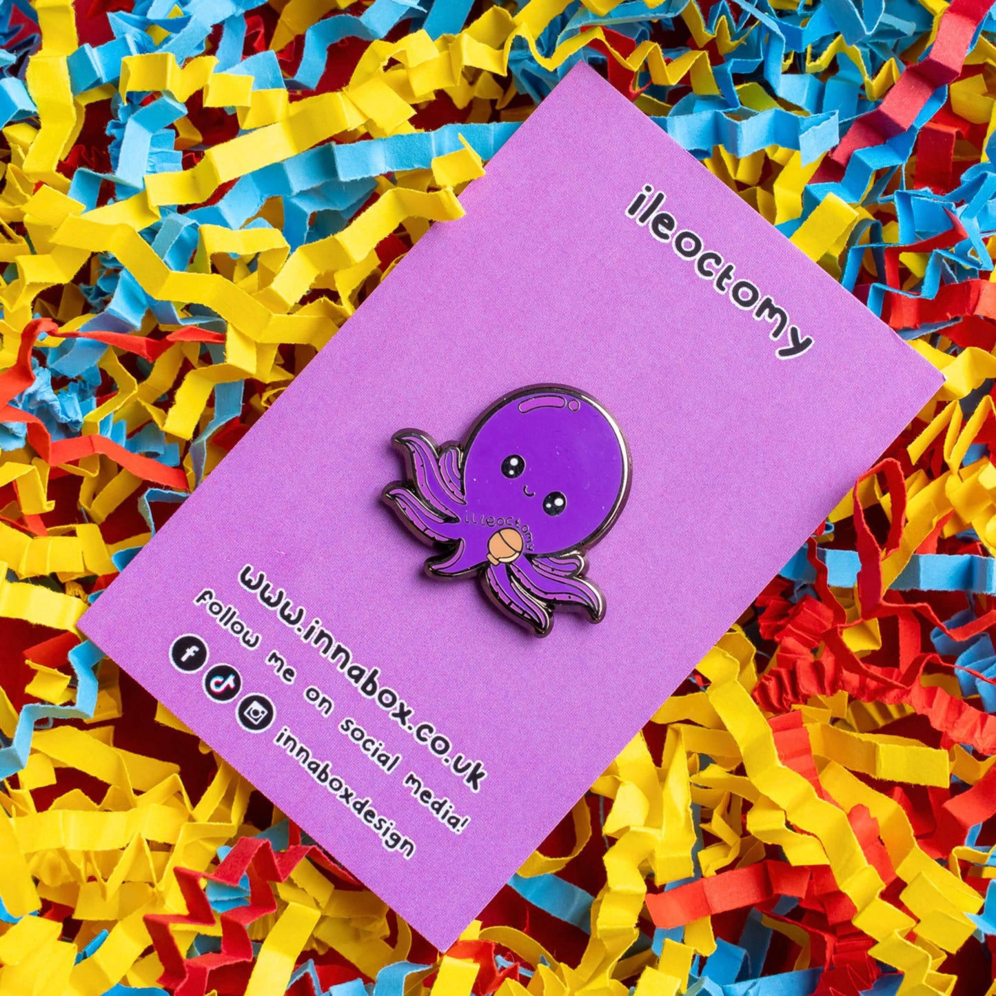 Ileoctomy Enamel Pin - Ileostomy on purple backing card laid on a yellow, blue and red card confetti background. The enamel pin is a cute smiling purple octopus sticker with text saying ileoctomy on its belly with a stoma bag underneath. Enamel pin designed to raise awareness for Ileostomy.