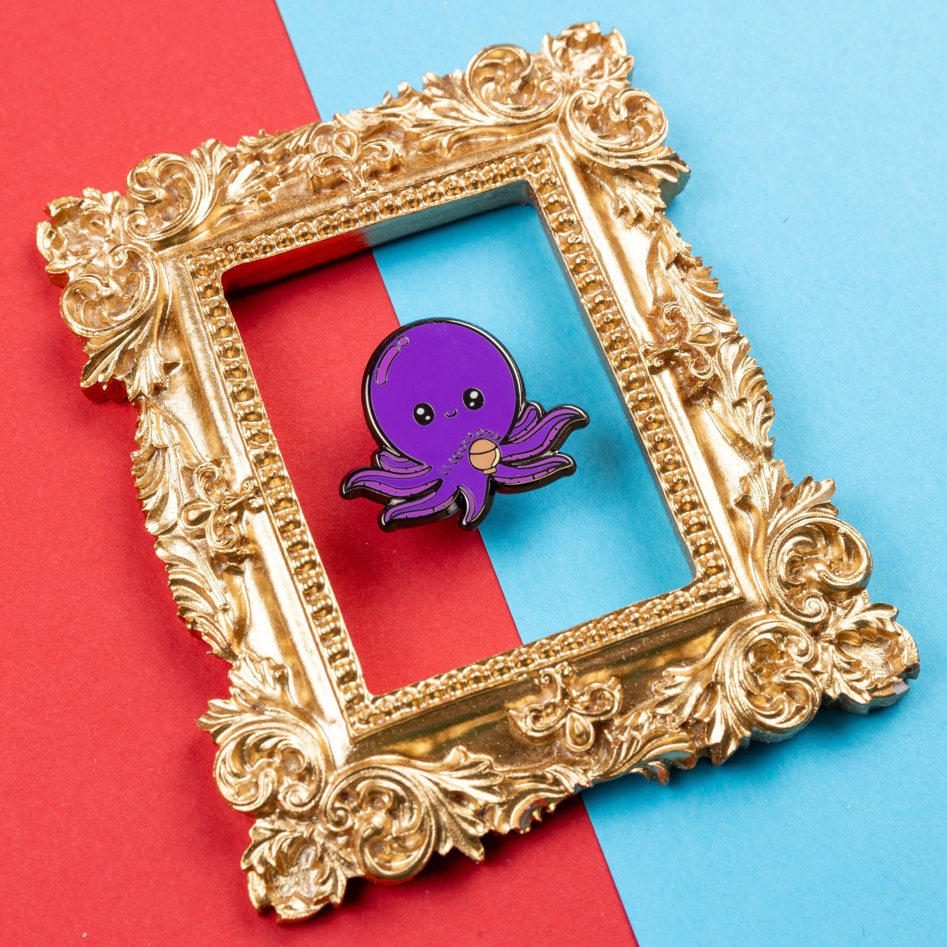 Ileoctomy Enamel Pin - Ileostomy on a blue and red card background inside a gold ornate frame. The enamel pin is a cute smiling purple octopus sticker with text saying ileoctomy on its belly with a stoma bag underneath. Enamel pin designed to raise awareness for Ileostomy.