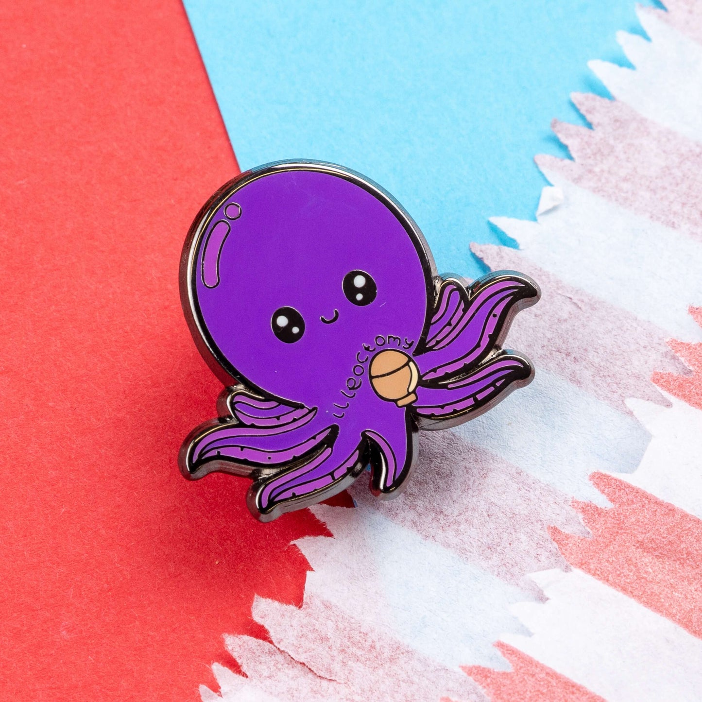 Ileoctomy Enamel Pin - Ileostomy on a blue and red card background. The enamel pin is a cute smiling purple octopus sticker with text saying ileoctomy on its belly with a stoma bag underneath. Enamel pin designed to raise awareness for Ileostomy.