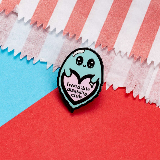 Invisible Disability Club Ghost Enamel Pin shown on a red and blue card background. The enamel pin is of a cute smiling ghost holding a pink heart with text saying invisible disability club. The enamel pin is designed to raise awareness for hidden and chronic disabilities.