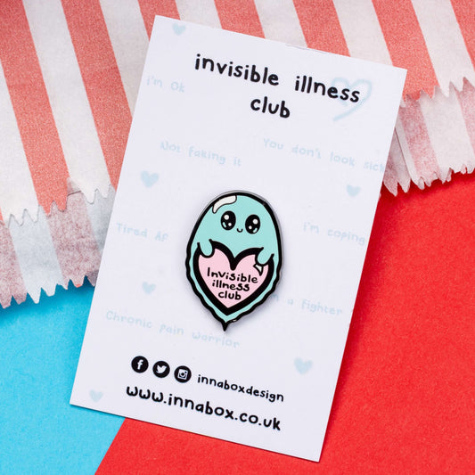 Invisible Illness Club Enamel Pin on white backing card laid on a red and blue card background. The enamel pin is of a cute smiling ghost holding a pink heart with text saying invisible illness club. The enamel pin is designed to raise awareness for hidden and chronic illnesses.