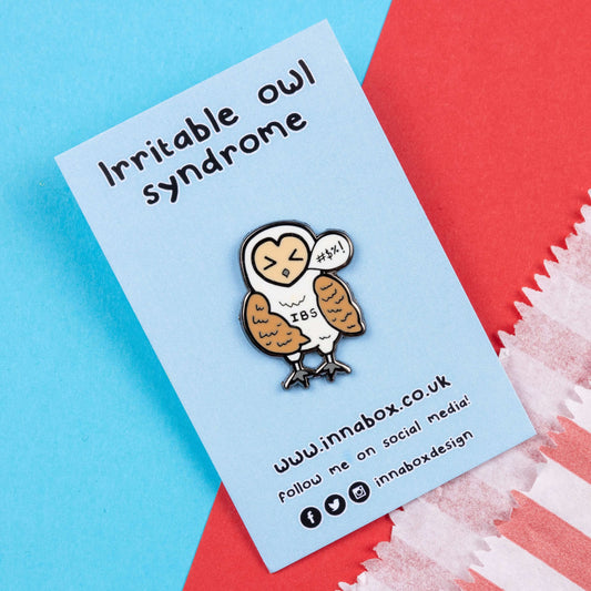 Irritable Owl Syndrome Enamel Pin - Irritable bowel syndrome (IBS) on pastel blue backing card on a red and blue background. The enamel pin is of a barn owl with a sweary text speech bubble and has its eyes shut with the letters IBS written on its belly. Hand drawn design to raise awareness for Irritable bowel syndrome (IBS).