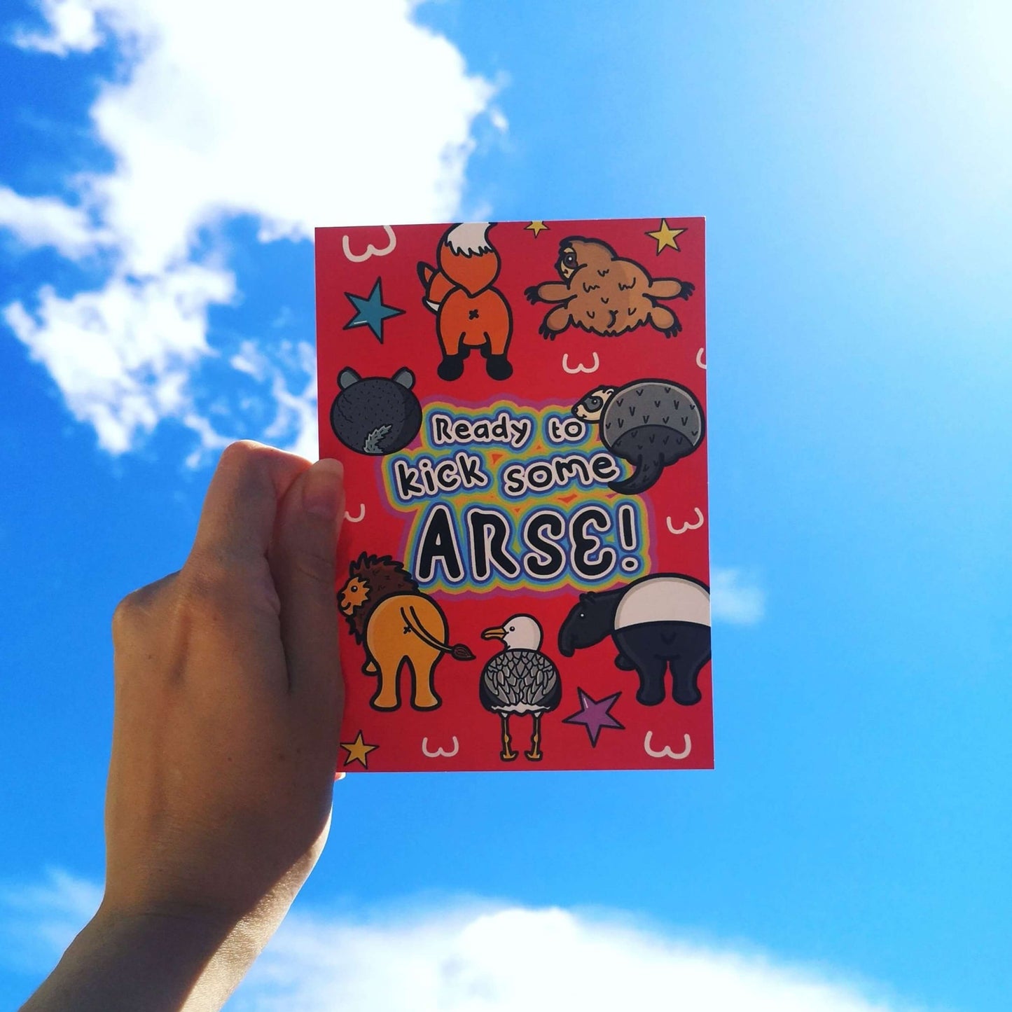 The Ready To Kick Some Arse Postcard held up to a blue sunny sky. The red card print features various animal bums (fox, sloth, chinchilla, badger, lion, seagull and tapir) with white bum outlines, yellow, blue and pink stars and centre rainbow outline text reading 'Ready to kick some arse!'.