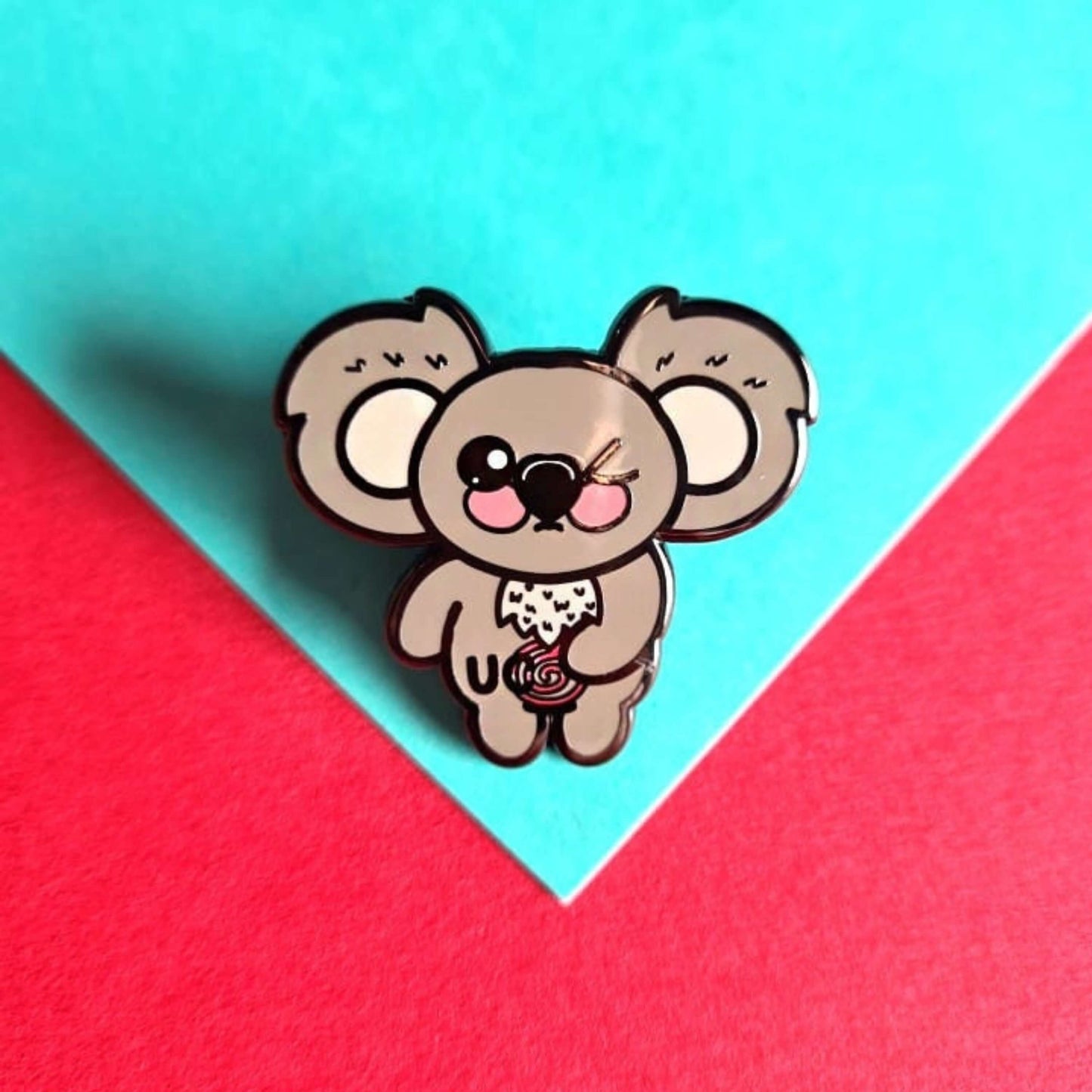 The Ulcerative Koalitis Koala Enamel Pin - Ulcerative Colitis UC on a red and blue background. The grey koala shaped enamel pin has one eye shut clutching its red swirly tummy with black text over it reading 'UC'. The hand drawn design is raising awareness for Ulcerative Colitis UC.