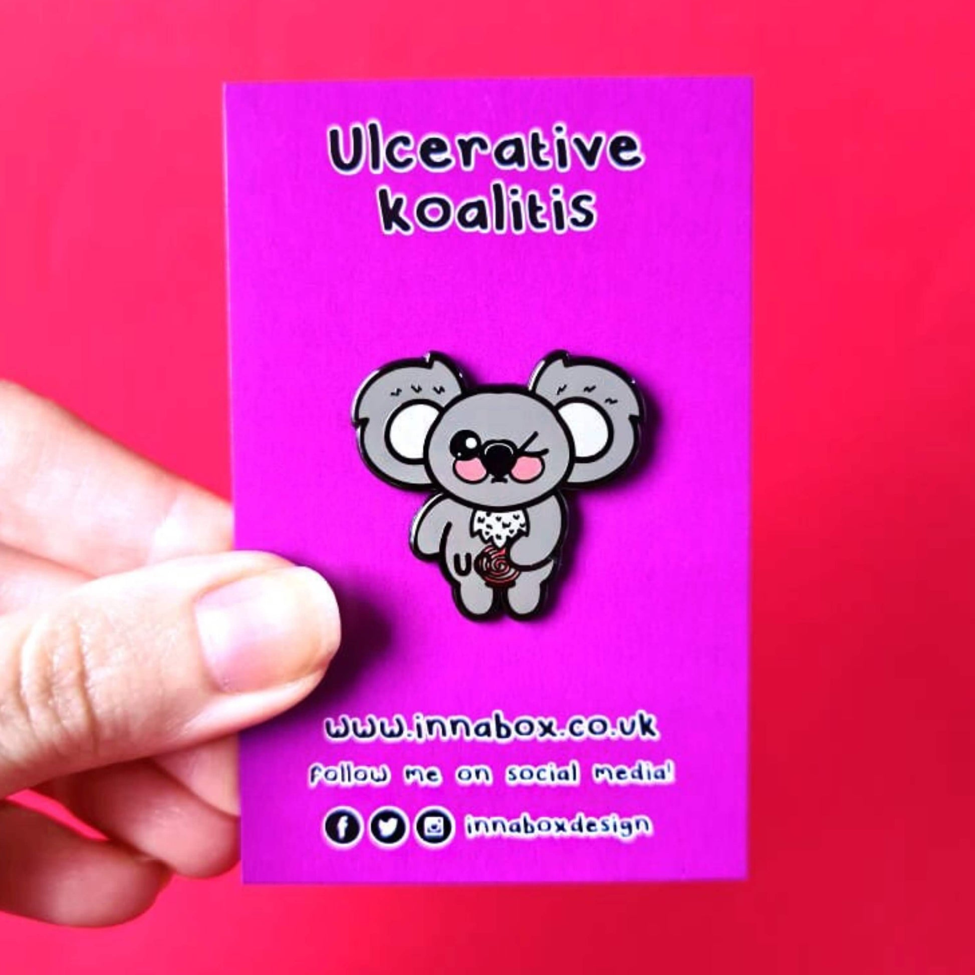 The Ulcerative Koalitis Koala Enamel Pin - Ulcerative Colitis UC on pink backing card held over a red background. The grey koala shaped enamel pin has one eye shut clutching its red swirly tummy with black text over it reading 'UC'. The hand drawn design is raising awareness for Ulcerative Colitis UC.