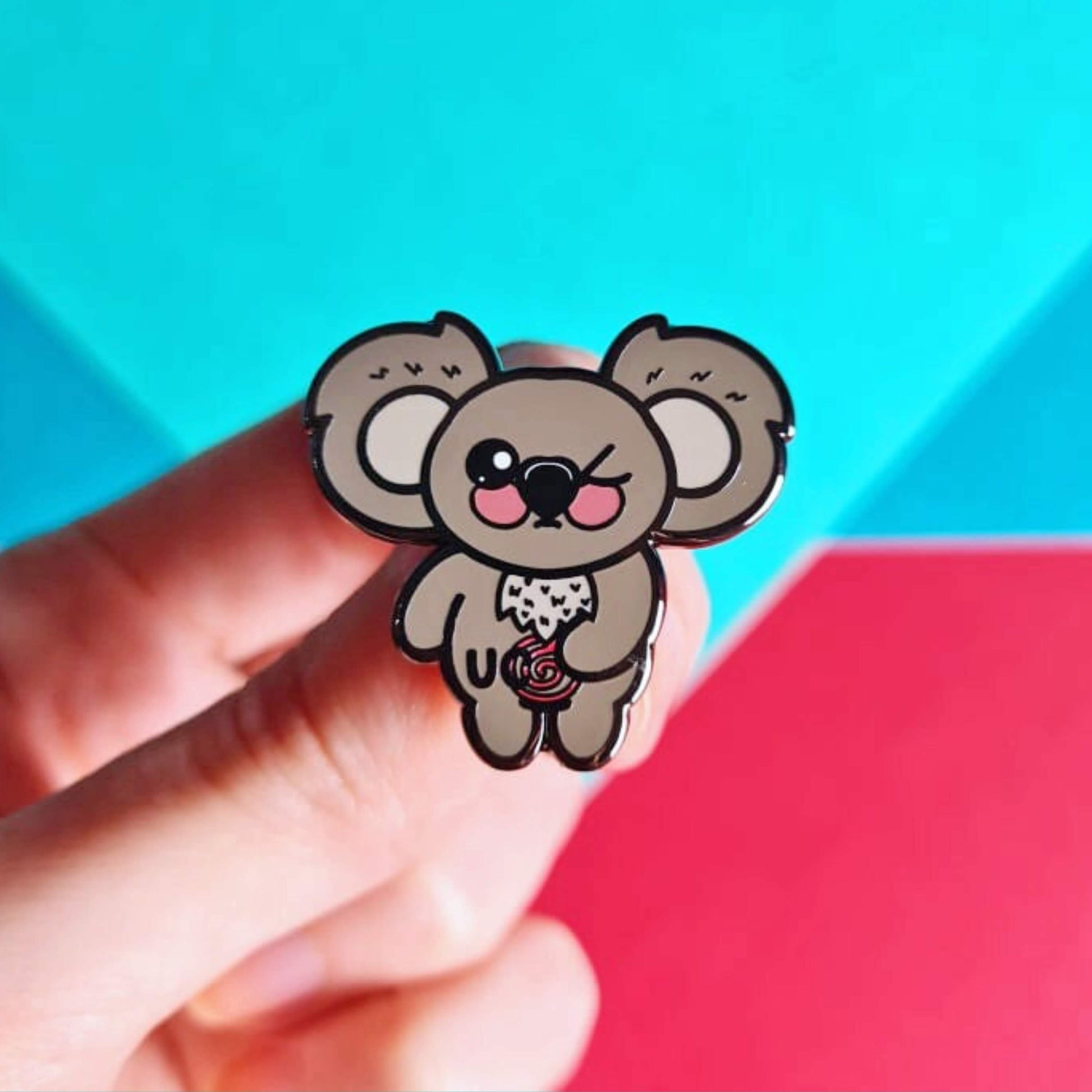 The Ulcerative Koalitis Koala Enamel Pin - Ulcerative Colitis UC held over a red and blue background. The grey koala shaped enamel pin has one eye shut clutching its red swirly tummy with black text over it reading 'UC'. The hand drawn design is raising awareness for Ulcerative Colitis UC.
