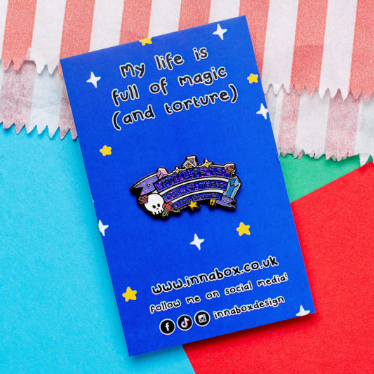 The My Life is Full of Magic & Torture Glitter Enamel Pin on blue backing card laid on a red, blue and green card background. The glittery finish enamel pin badge features a purple banner sash of black text reading 'my life is full of magic (and torture)' with yellow sparkles, crystals, red roses, tarot cards and a skull head.