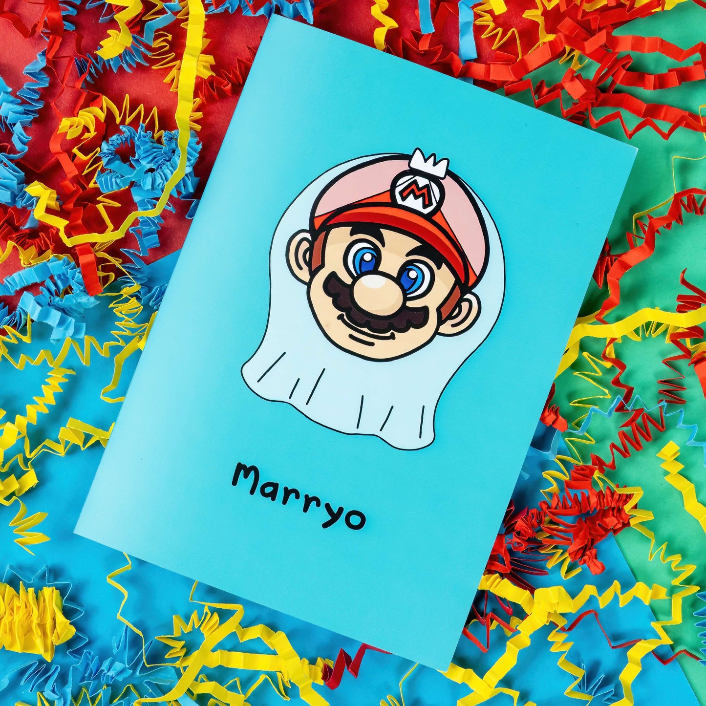 The Marryo - Mario Engagement Wedding Card on a red, blue and green background with red, yellow and blue crinkle card confetti. The blue a6 congratulations card features a smiling nintendo mario character head wearing a white bridal wedding veil, underneath him is black text reading 'Marryo'.