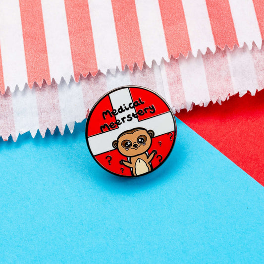 Medical Mystery Meerkat Enamel Pin on a red and blue background. The enamel pin is a red circle with white first aid cross and a cute meerkat at the bottom with big eyes and has it's little arms open wide. There are black question marks around the meerkat and 'medical meerstery' written above it in black writing. Hand drawn design made to raise awareness for invisible and chronic illnesses.