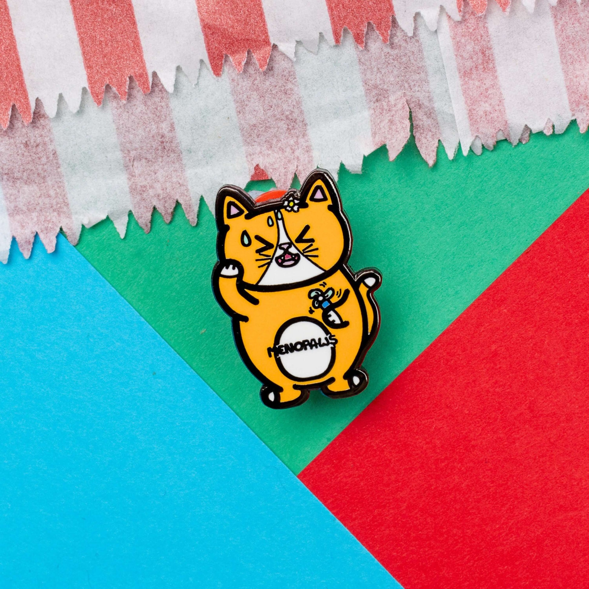 Menopaws Enamel Pin - Menopause on a red, blue and green card background. The enamel pin is of a menopausal ginger and white cat with a daisy in it's hair. The cat is holding an electric fan and is looking hot with sweat droplets on it's face. Hand drawn design made to raise awareness for menopause.