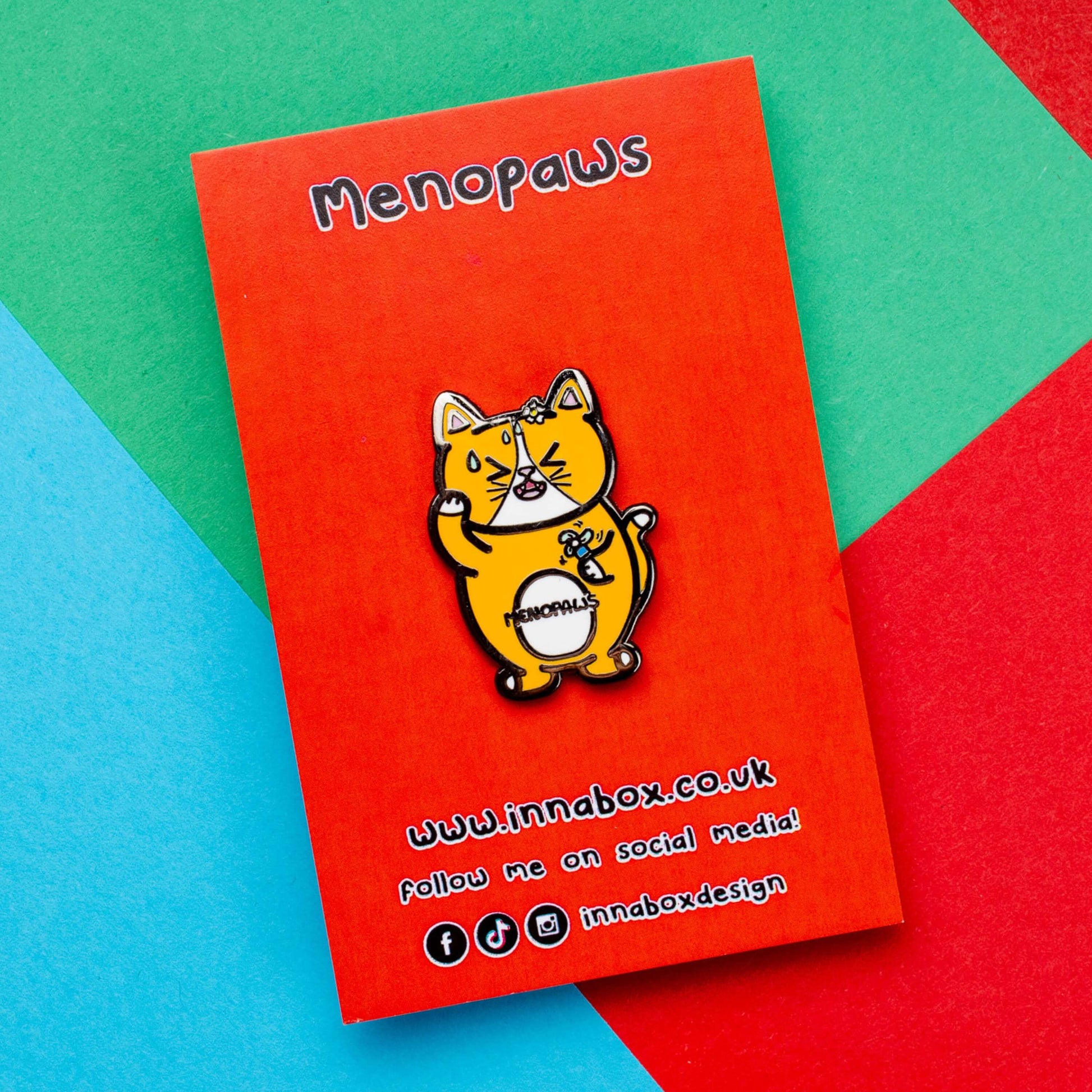 Menopaws Enamel Pin - Menopause on red backing card laid on a red, blue and green card background. The enamel pin is of a menopausal ginger and white cat with a daisy in it's hair. The cat is holding an electric fan and is looking hot with sweat droplets on it's face. Hand drawn design made to raise awareness for menopause.