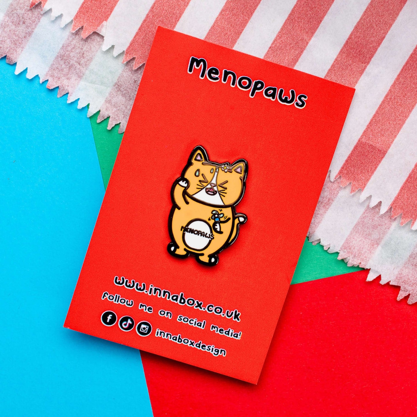 Menopaws Enamel Pin - Menopause on red backing card laid on a red, blue and green card background. The enamel pin is of a menopausal ginger and white cat with a daisy in it's hair. The cat is holding an electric fan and is looking hot with sweat droplets on it's face. Hand drawn design made to raise awareness for menopause.