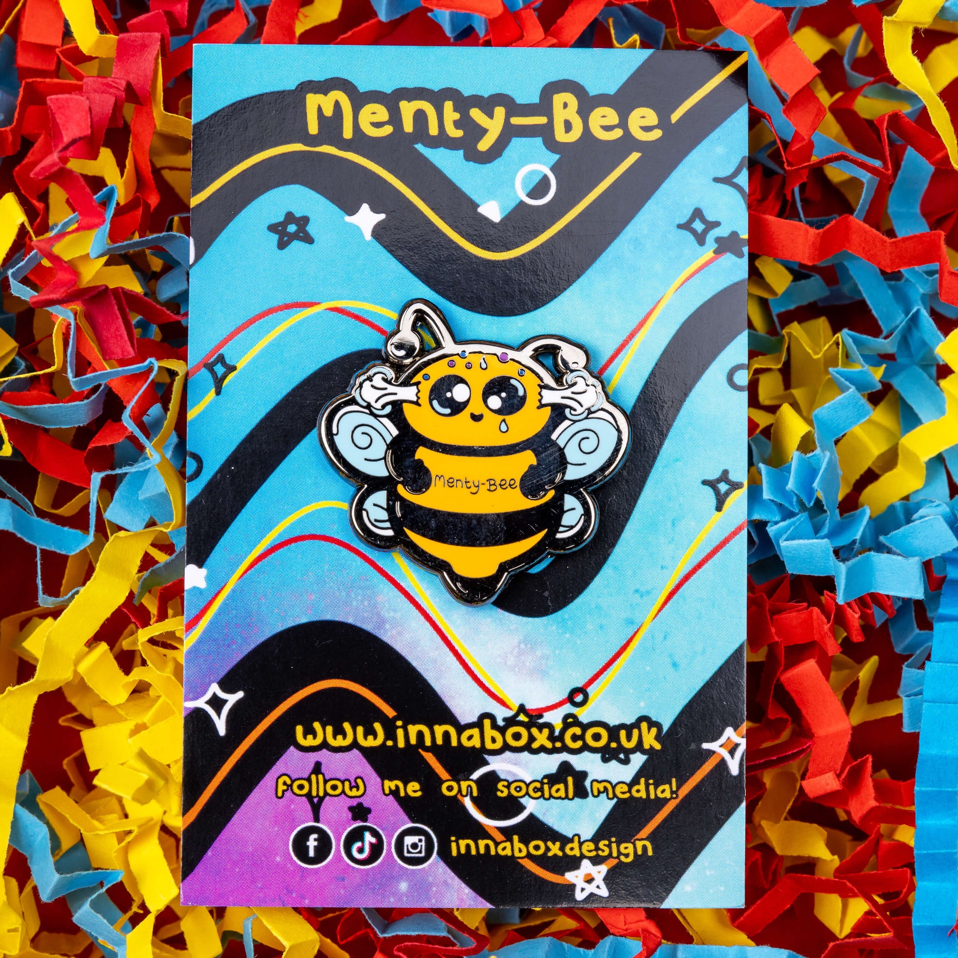 Menty-Bee Enamel Pin - Mental Breakdown pinned to its backing card with card confetti in the background. The enamel pin is of a bee looking stressed with steam coming out of its head and the text menty-Bee on its chest. Hand drawn design made to raise awareness for mental breakdowns