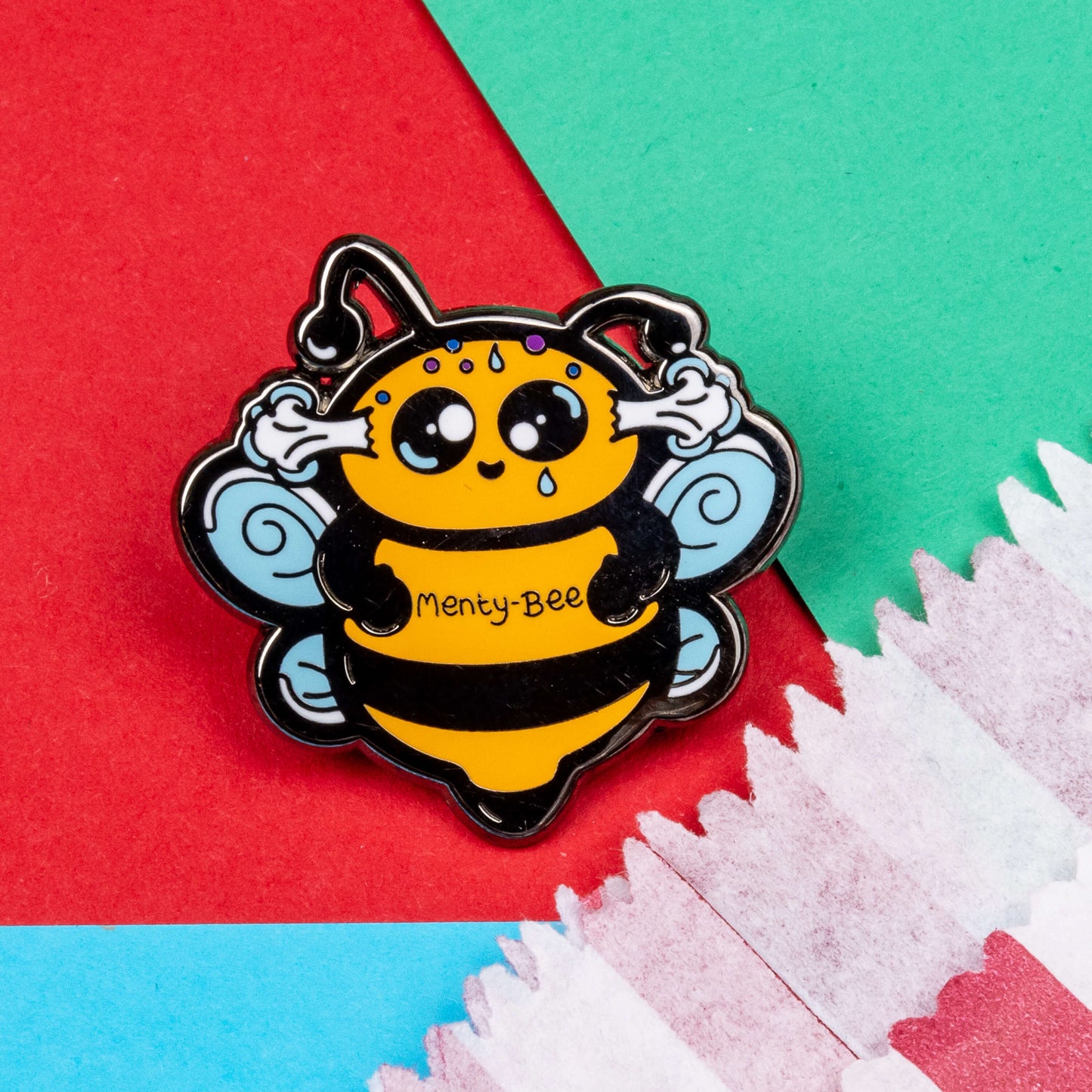 Menty-Bee Enamel Pin - Mental Breakdown on a red and blue background next to a red and white striped paper bag. The enamel pin is of a bee looking stressed with steam coming out of its head and the text menty-Bee on its chest. Hand drawn design made to raise awareness for mental breakdowns