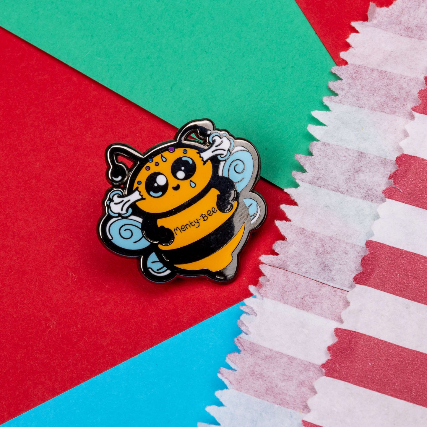 Menty-Bee Enamel Pin - Mental Breakdown on a red and blue background next to a red and white striped paper bag. The enamel pin is of a bee looking stressed with steam coming out of its head and the text menty-Bee on its chest. Hand drawn design made to raise awareness for mental breakdowns