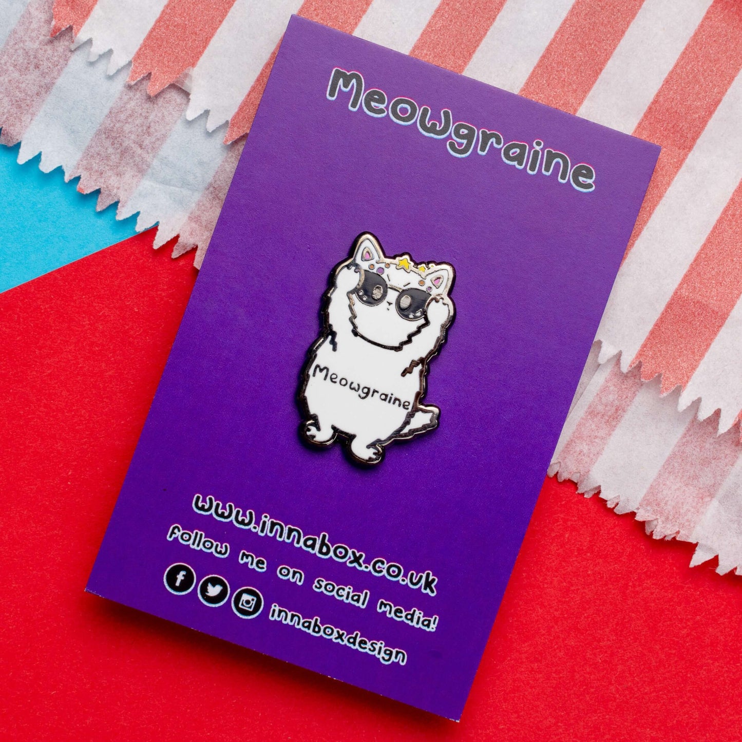 The Meowgraine Cat Enamel Pin - Migraine on purple backing card laid on a red and blue background. A white stressed cat clutching a pair of black sunglasses to its eyes with multicoloured spots and stars over its head, across its middle reads 'meowgraine'. The hand drawn design is raising awareness for migraines and headaches.