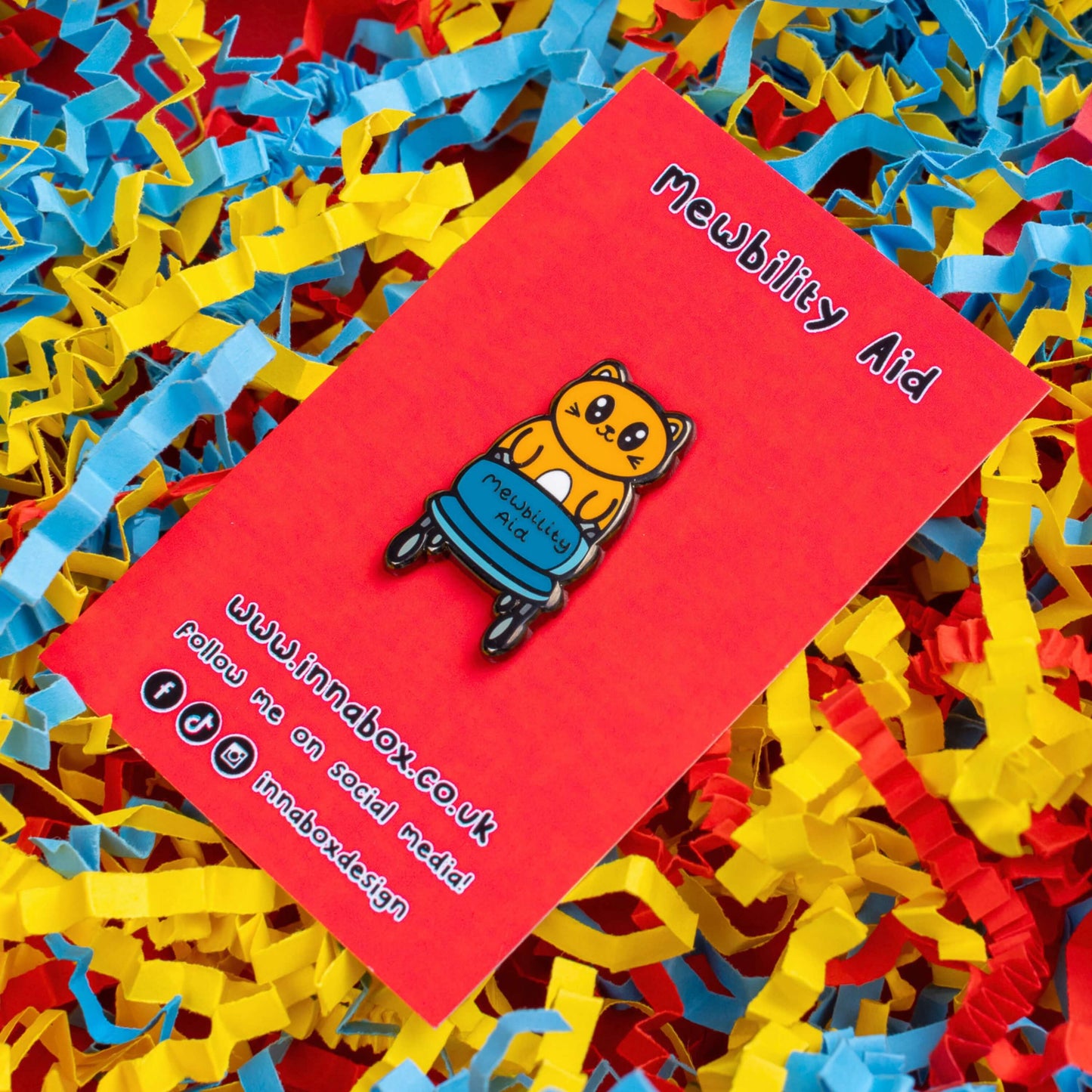 The Mewbility Aid Cat Enamel Pin - Mobility Aid on red backing card laid on red, yellow and blue card confetti background. An orange smiling cat walking upright with a blue mobility aid, across the walking chair reads 'mewbility aid'. The hand drawn designs is raising awareness for mobility aids.