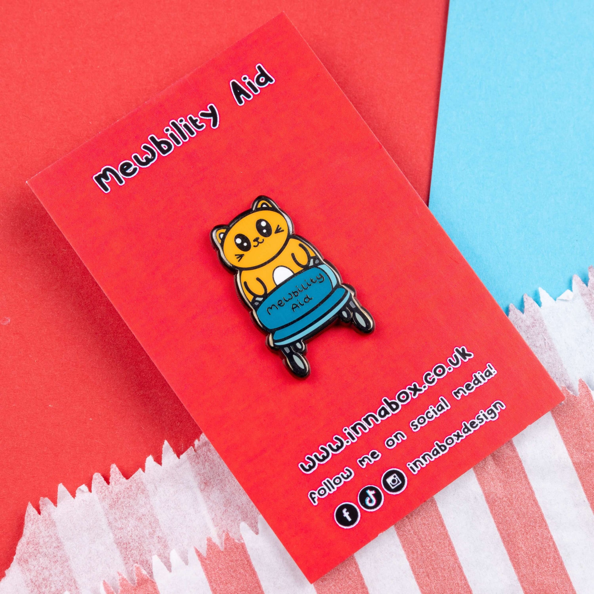 The Mewbility Aid Cat Enamel Pin - Mobility Aid on red backing card on red and blue card. An orange smiling cat walking upright with a blue mobility aid, across the walking chair reads 'mewbility aid'. The hand drawn designs is raising awareness for mobility aids.