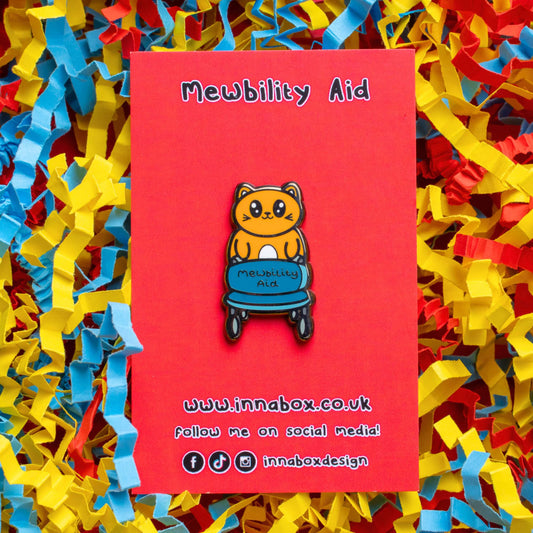 The Mewbility Aid Cat Enamel Pin - Mobility Aid on red backing card laid on red, yellow and blue card confetti background. An orange smiling cat walking upright with a blue mobility aid, across the walking chair reads 'mewbility aid'. The hand drawn designs is raising awareness for mobility aids.