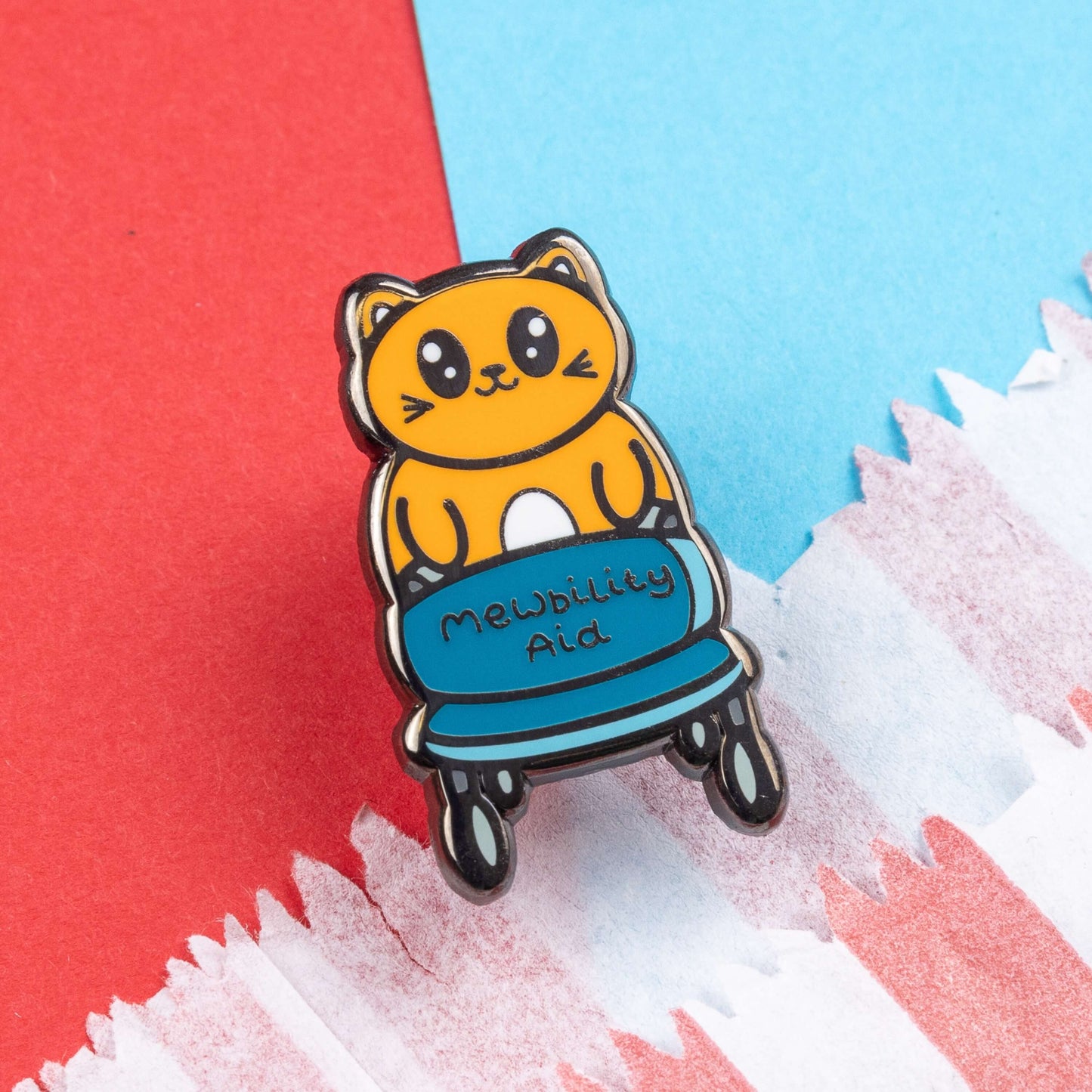 The Mewbility Aid Cat Enamel Pin - Mobility Aid on red and blue card. An orange smiling cat walking upright with a blue mobility aid, across the walking chair reads 'mewbility aid'. The hand drawn designs is raising awareness for mobility aids.