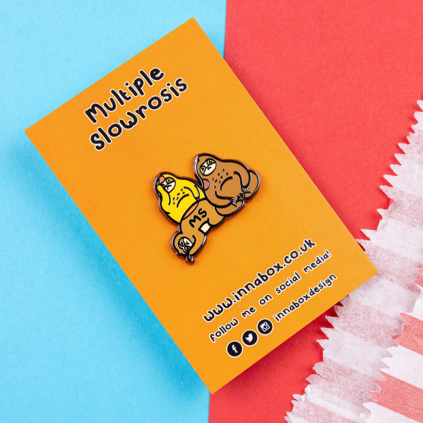 The Multiple Slowrosis Sloth Enamel Pin - Multiple Sclerosis / MS on orange backing card laid on a red and blue background. The pin of three sad sloths, two brown and one yellow, with red patches all over their body. Across one in black text reads 'MS'. The hand drawn design is raising awareness for Multiple Sclerosis / MS.