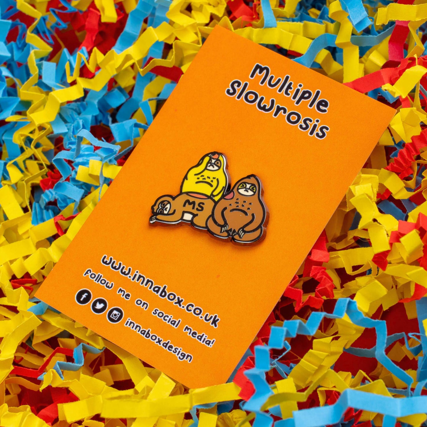 The Multiple Slowrosis Sloth Enamel Pin - Multiple Sclerosis / MS on orange backing card laid on a red, yellow and blue card confetti background. The pin of three sad sloths, two brown and one yellow, with red patches all over their body. Across one in black text reads 'MS'. The hand drawn design is raising awareness for Multiple Sclerosis / MS.