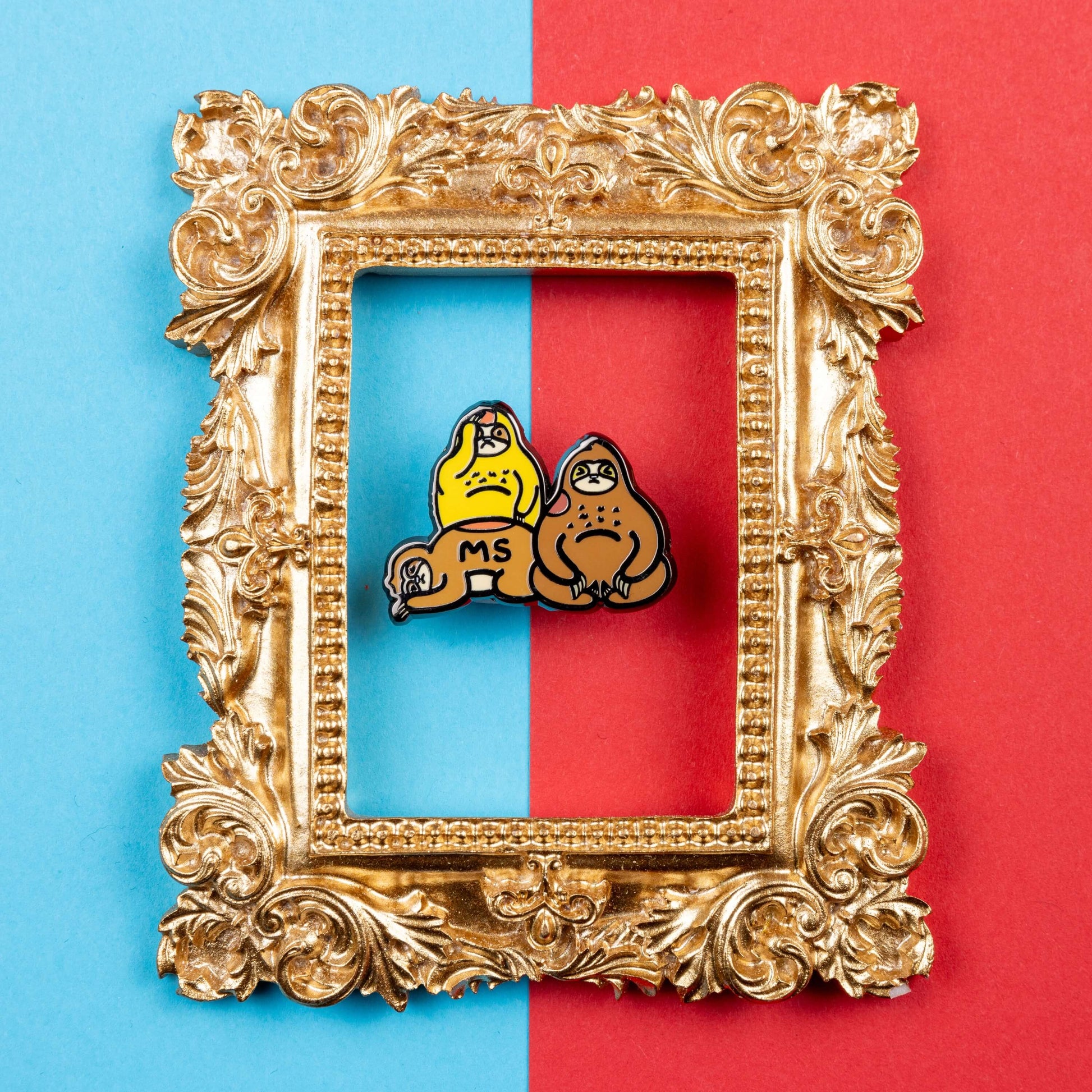 The Multiple Slowrosis Sloth Enamel Pin - Multiple Sclerosis / MS on a blue and red background inside a gold ornate frame. The pin of three sad sloths, two brown and one yellow, with red patches all over their body. Across one in black text reads 'MS'. The hand drawn design is raising awareness for Multiple Sclerosis / MS.