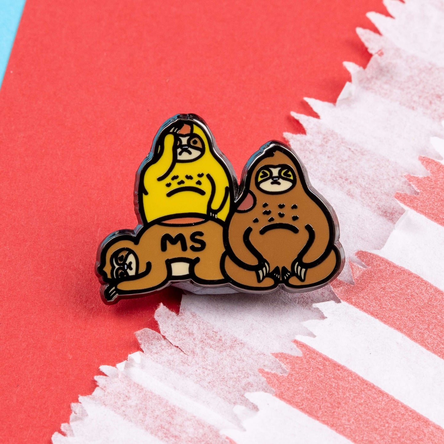 The Multiple Slowrosis Sloth Enamel Pin - Multiple Sclerosis / MS on a red and blue background. The pin of three sad sloths, two brown and one yellow, with red patches all over their body. Across one in black text reads 'MS'. The hand drawn design is raising awareness for Multiple Sclerosis / MS.