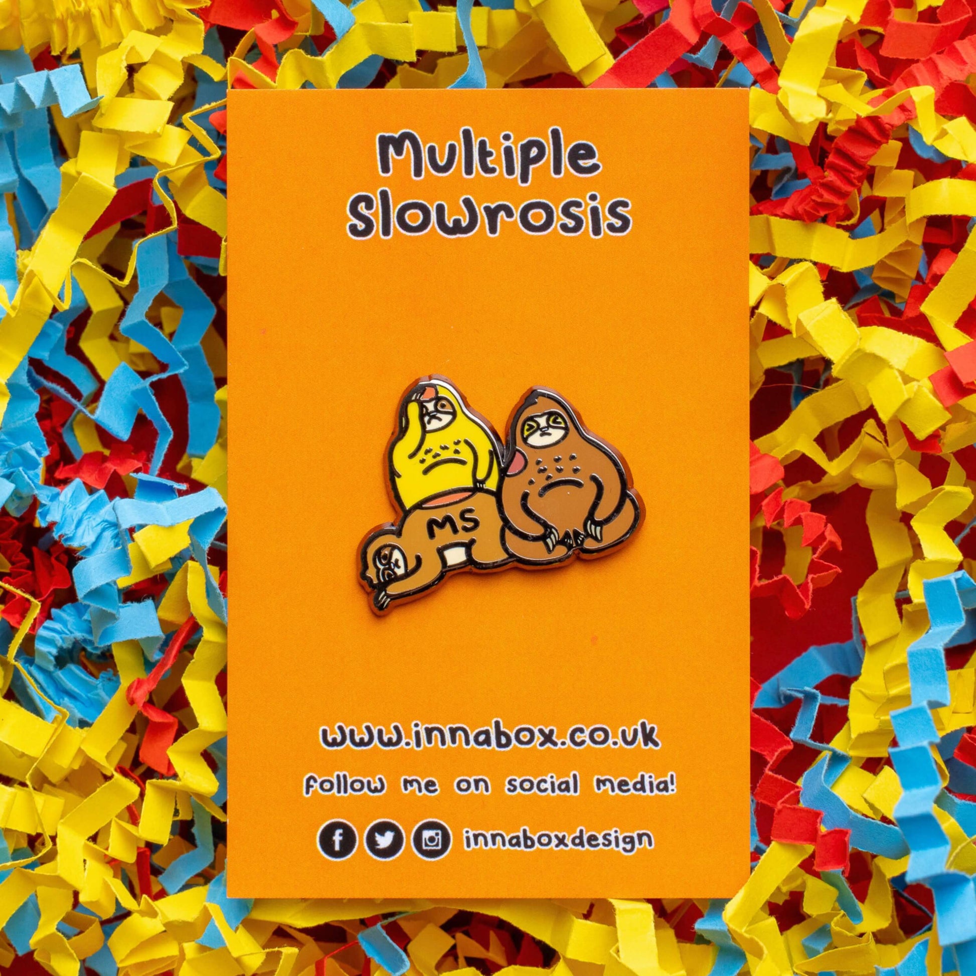 The Multiple Slowrosis Sloth Enamel Pin - Multiple Sclerosis / MS on orange backing card laid on a red, yellow and blue card confetti background. The pin of three sad sloths, two brown and one yellow, with red patches all over their body. Across one in black text reads 'MS'. The hand drawn design is raising awareness for Multiple Sclerosis / MS.