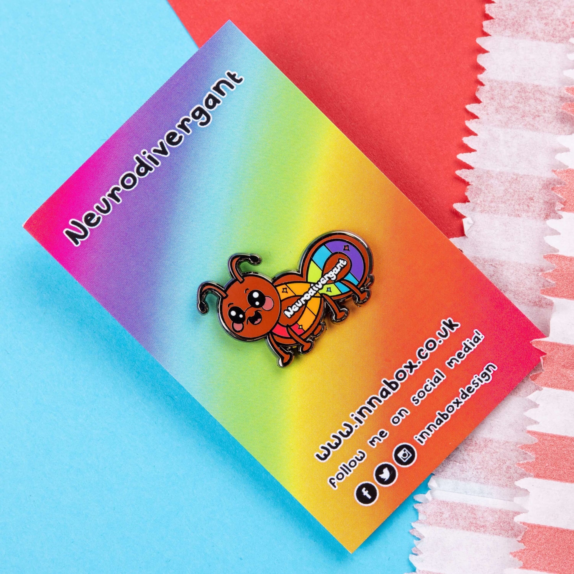 The Neurodivergant Ant Enamel Pin - Neurodivergent on rainbow backing card with innabox social media handles along the bottom laid on a blue and red background. The brown ant shape enamel pin is smiling with a rainbow infinity symbol and white text reading 'neurodivergant' with sparkles across its body. The hand drawn design is raising awareness for neurodiversity.