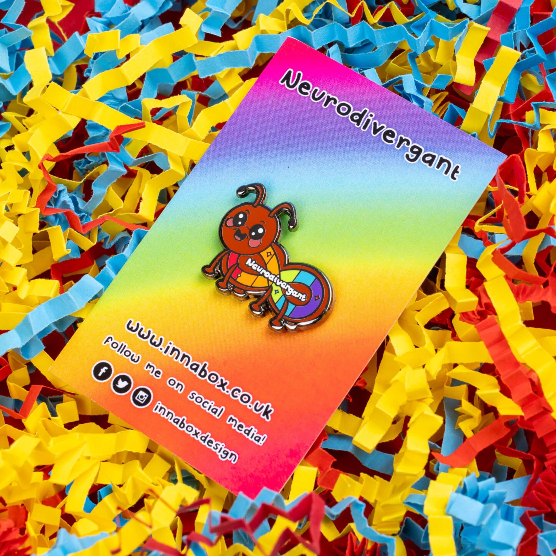 The Neurodivergant Ant Enamel Pin - Neurodivergent on rainbow backing card with innabox social media handles along the bottom laid on a blue, yellow and red card confetti background. The brown ant shape enamel pin is smiling with a rainbow infinity symbol and white text reading 'neurodivergant' with sparkles across its body. The hand drawn design is raising awareness for neurodiversity.