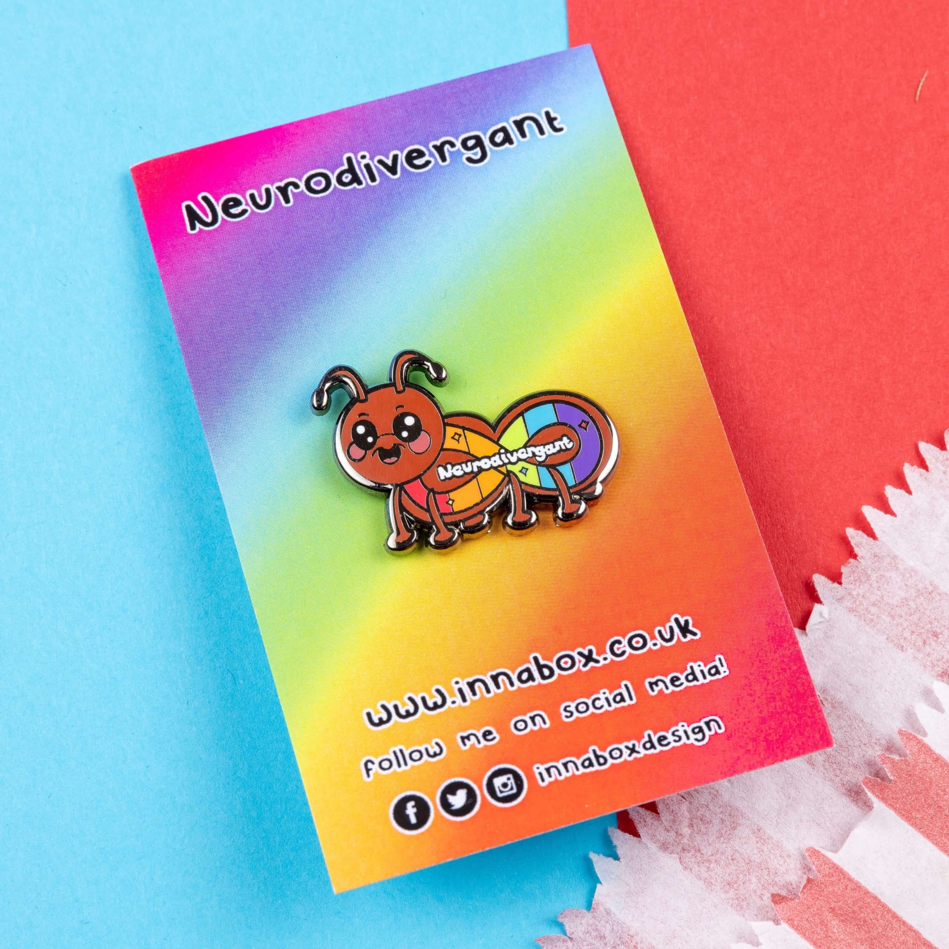 The Neurodivergant Ant Enamel Pin - Neurodivergent on rainbow backing card with innabox social media handles along the bottom laid on a blue and red background. The brown ant shape enamel pin is smiling with a rainbow infinity symbol and white text reading 'neurodivergant' with sparkles across its body. The hand drawn design is raising awareness for neurodiversity.