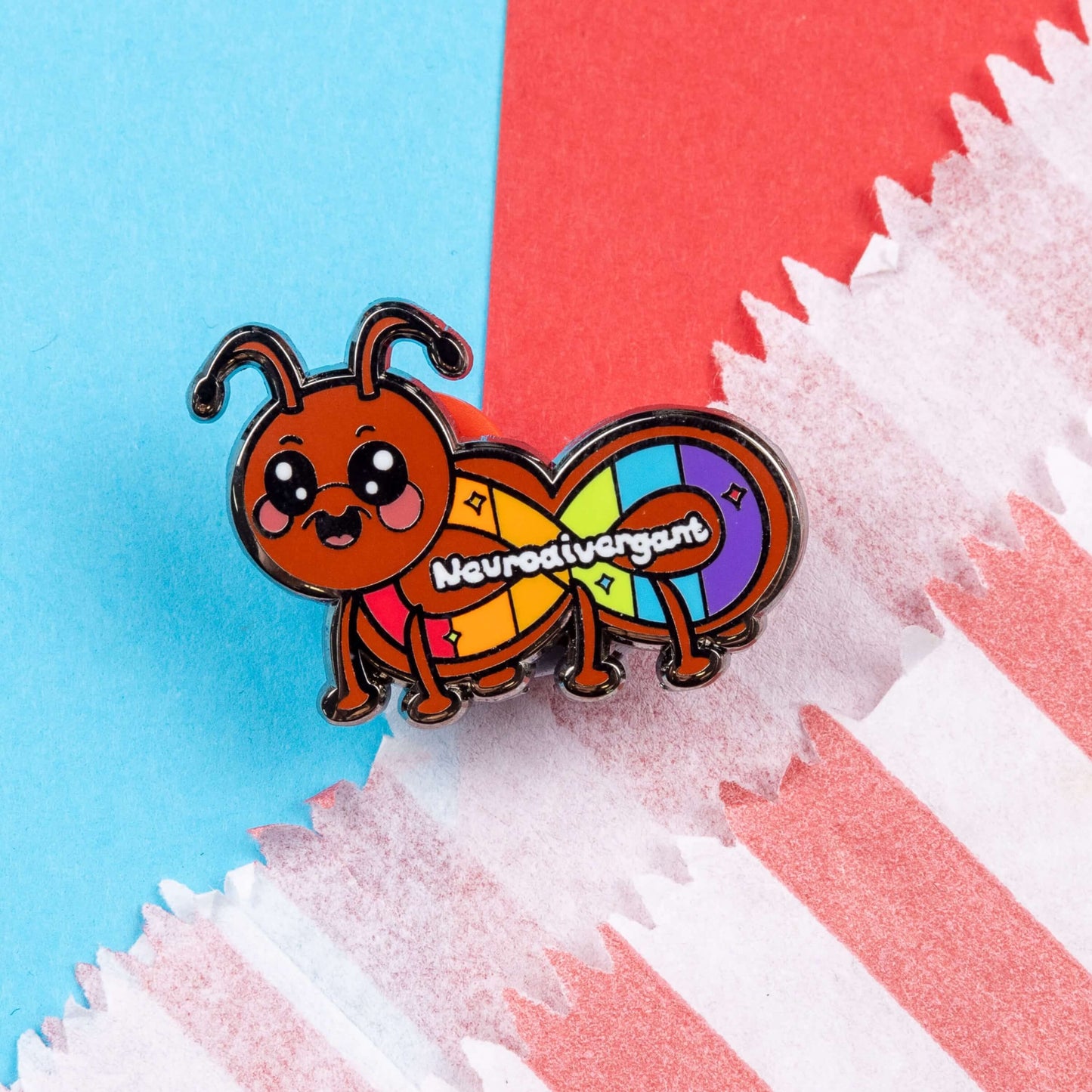 The Neurodivergant Ant Enamel Pin - Neurodivergent on a red and blue background. The brown ant shape enamel pin is smiling with a rainbow infinity symbol and white text reading 'neurodivergant' with sparkles across its body. The hand drawn design is raising awareness for neurodiversity.