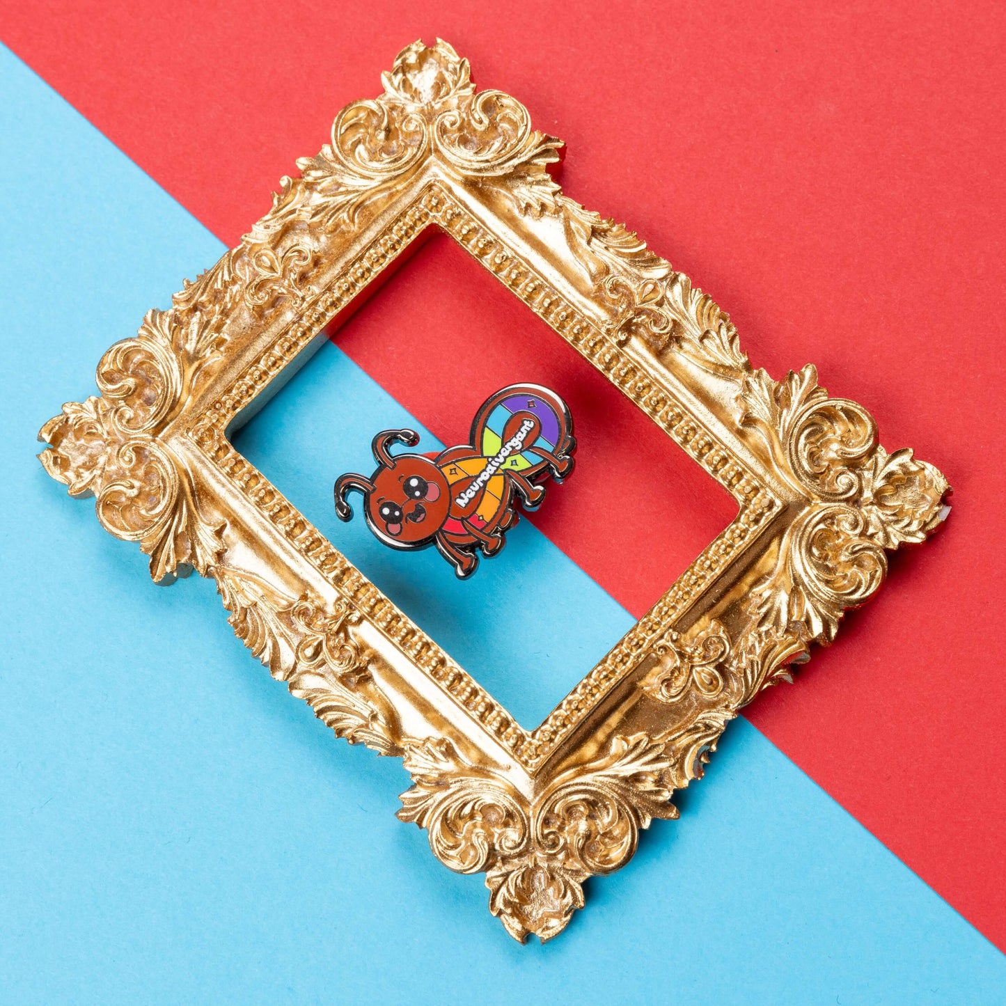 The Neurodivergant Ant Enamel Pin - Neurodivergent on a red and blue background inside a gold ornate frame. The brown ant shape enamel pin is smiling with a rainbow infinity symbol and white text reading 'neurodivergant' with sparkles across its body. The hand drawn design is raising awareness for neurodiversity.