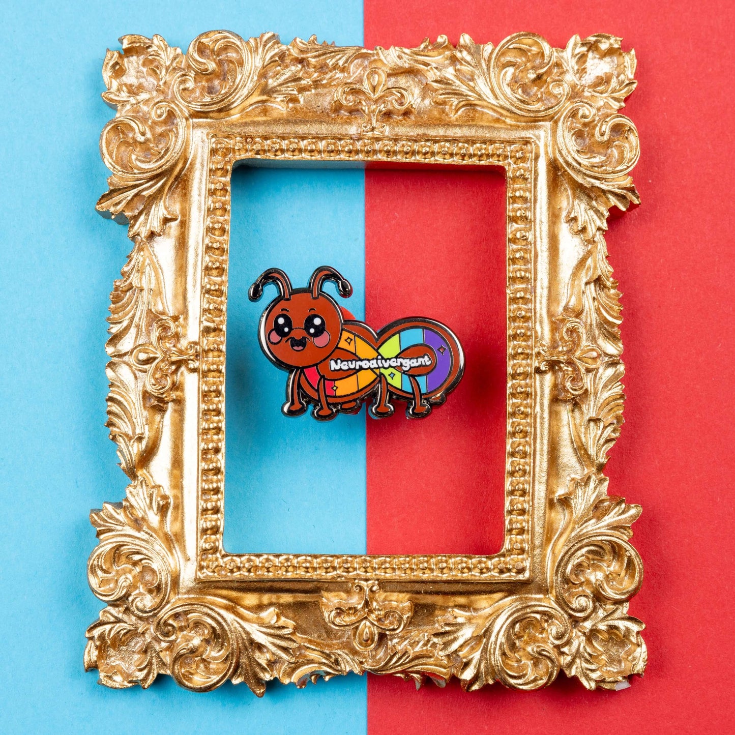 The Neurodivergant Ant Enamel Pin - Neurodivergent on a red and blue background inside a gold ornate frame. The brown ant shape enamel pin is smiling with a rainbow infinity symbol and white text reading 'neurodivergant' with sparkles across its body. The hand drawn design is raising awareness for neurodiversity.