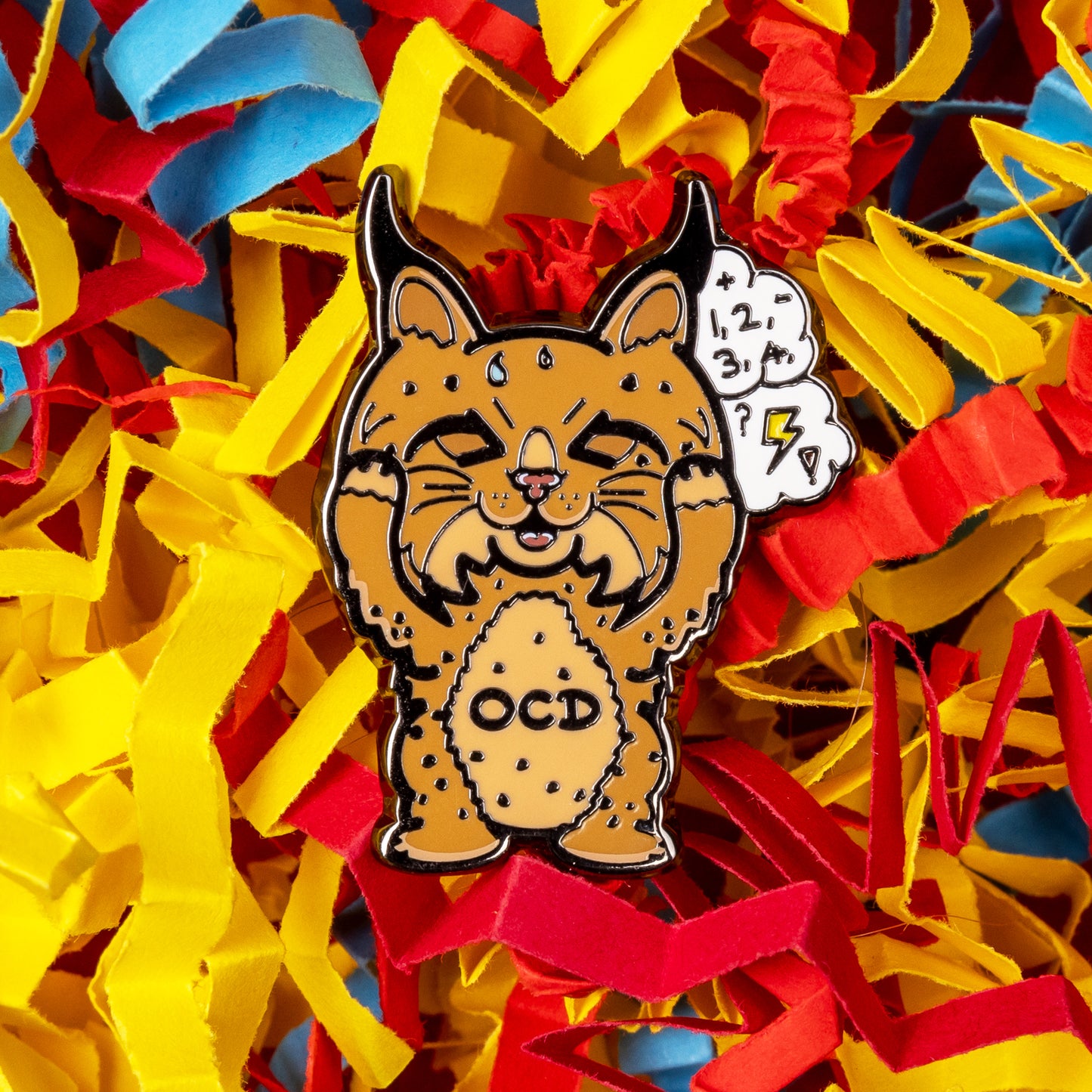 Bobsessive Compulsive Disorder Enamel Pin - OCD on a red, yellow and blue coloured card confetti background. The bobcat shape pin is brown with a stressed expression clutching its face with a thought bubble of numbers, question marks and lightning bolts. Across its tummy reads OCD. The pin was designed to raise awareness of obsessive compulsive disorder.