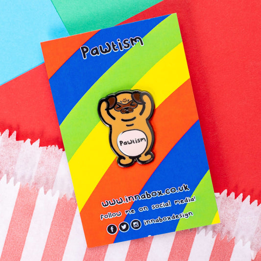 Pawtism Enamel Pin - Autism on rainbow stripe backing card laid on a red, green and blue card background. The enamel pin is of a brown pug with its hands on its ears and the word pawtism written across its belly. The enamel pin is to raise awareness for neurodiversity and autism.