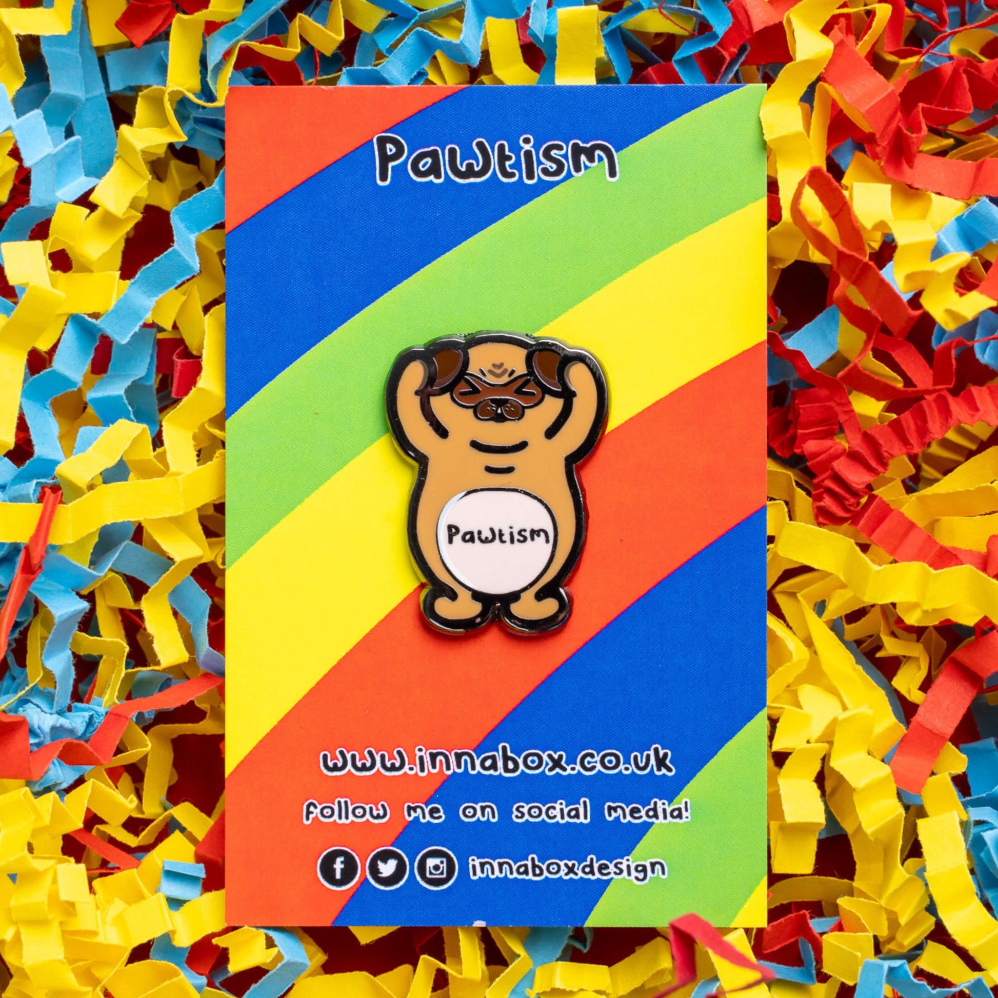 Pawtism Enamel Pin - Autism on rainbow stripe backing card laid on a red, yellow and blue card confetti background. The enamel pin is of a brown pug with its hands on its ears and the word pawtism written across its belly. The enamel pin is to raise awareness for neurodiversity and autism.