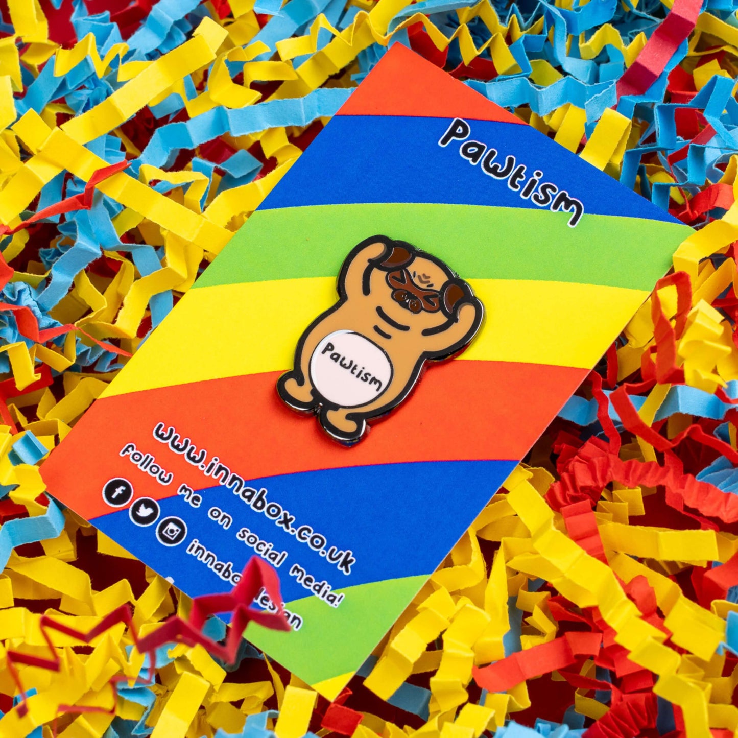 Pawtism Enamel Pin - Autism on rainbow stripe backing card laid on a red, yellow and blue card confetti background. The enamel pin is of a brown pug with its hands on its ears and the word pawtism written across its belly. The enamel pin is to raise awareness for neurodiversity and autism.